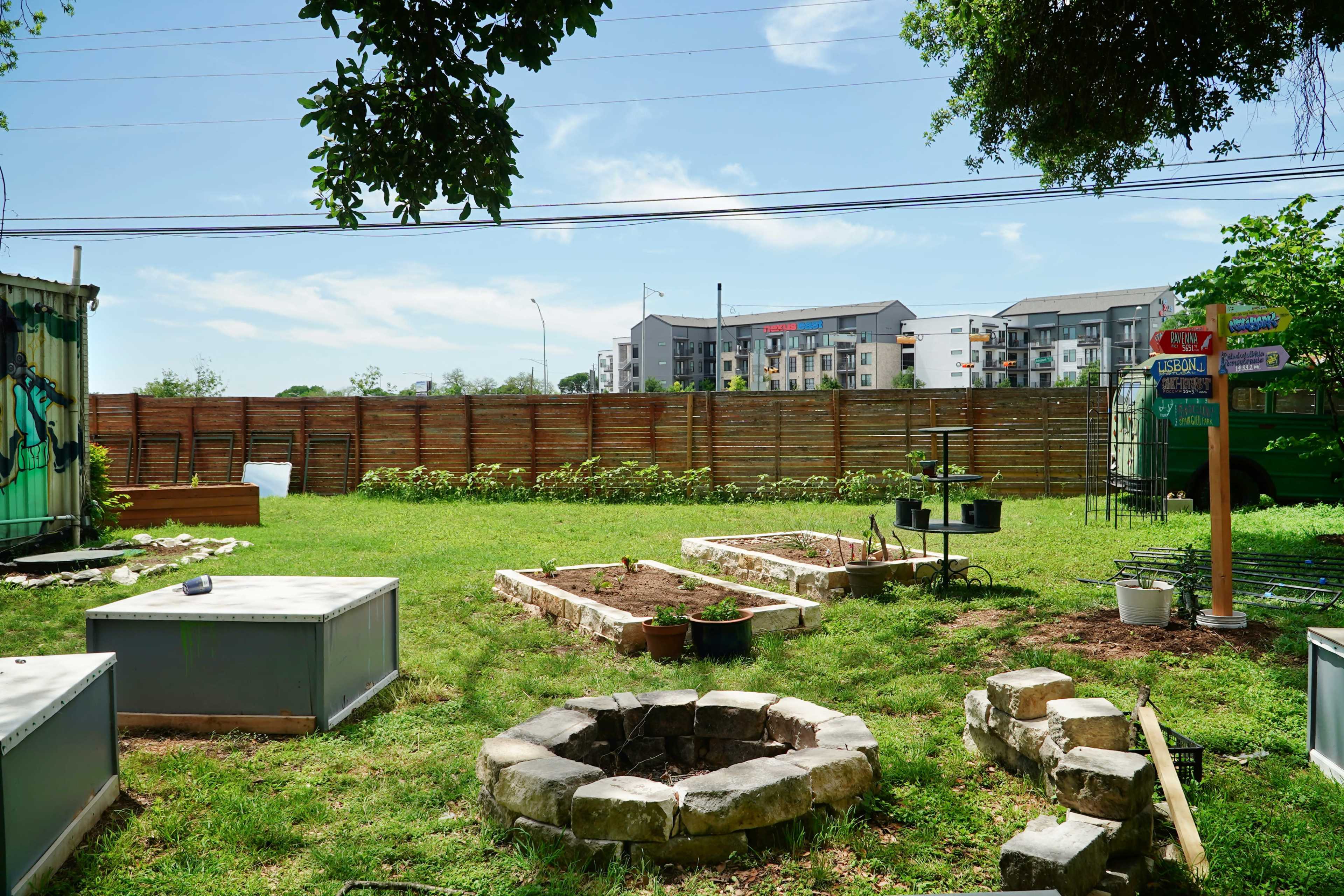 A grassy backyard with several raised garden beds, a stone fire pit, and a wooden fence in the background, along with apartment buildings beyond the fence.