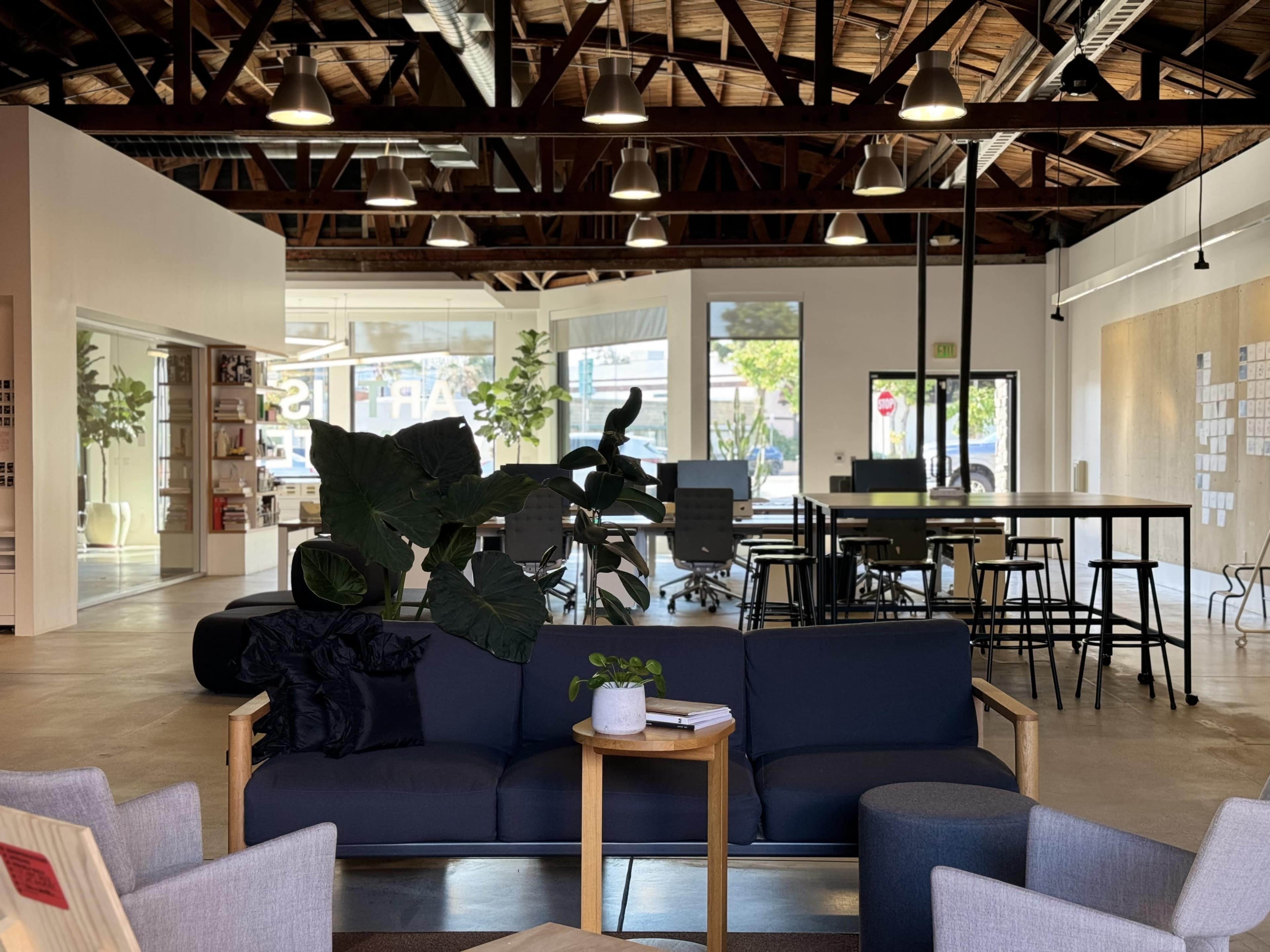 The image shows a spacious, modern office interior featuring seating areas with plants, a communal workspace with tables and chairs, and large windows allowing natural light.