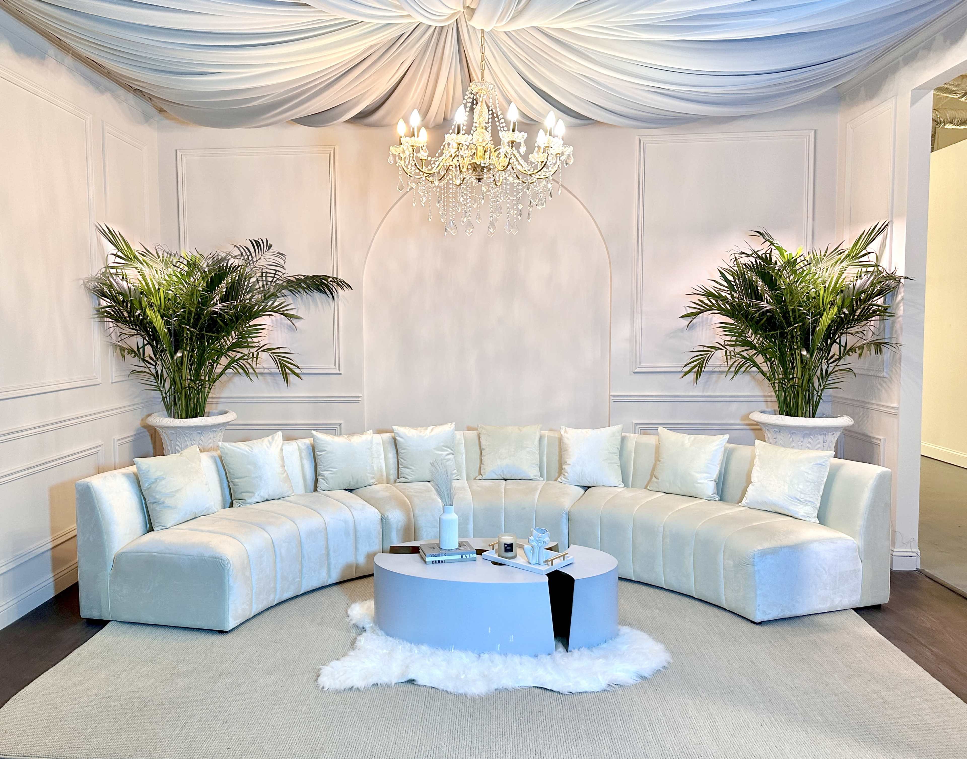 The image shows a spacious, elegantly decorated room featuring a curved white sofa, a round coffee table, two large potted plants, and a chandelier hanging from a draped ceiling.
