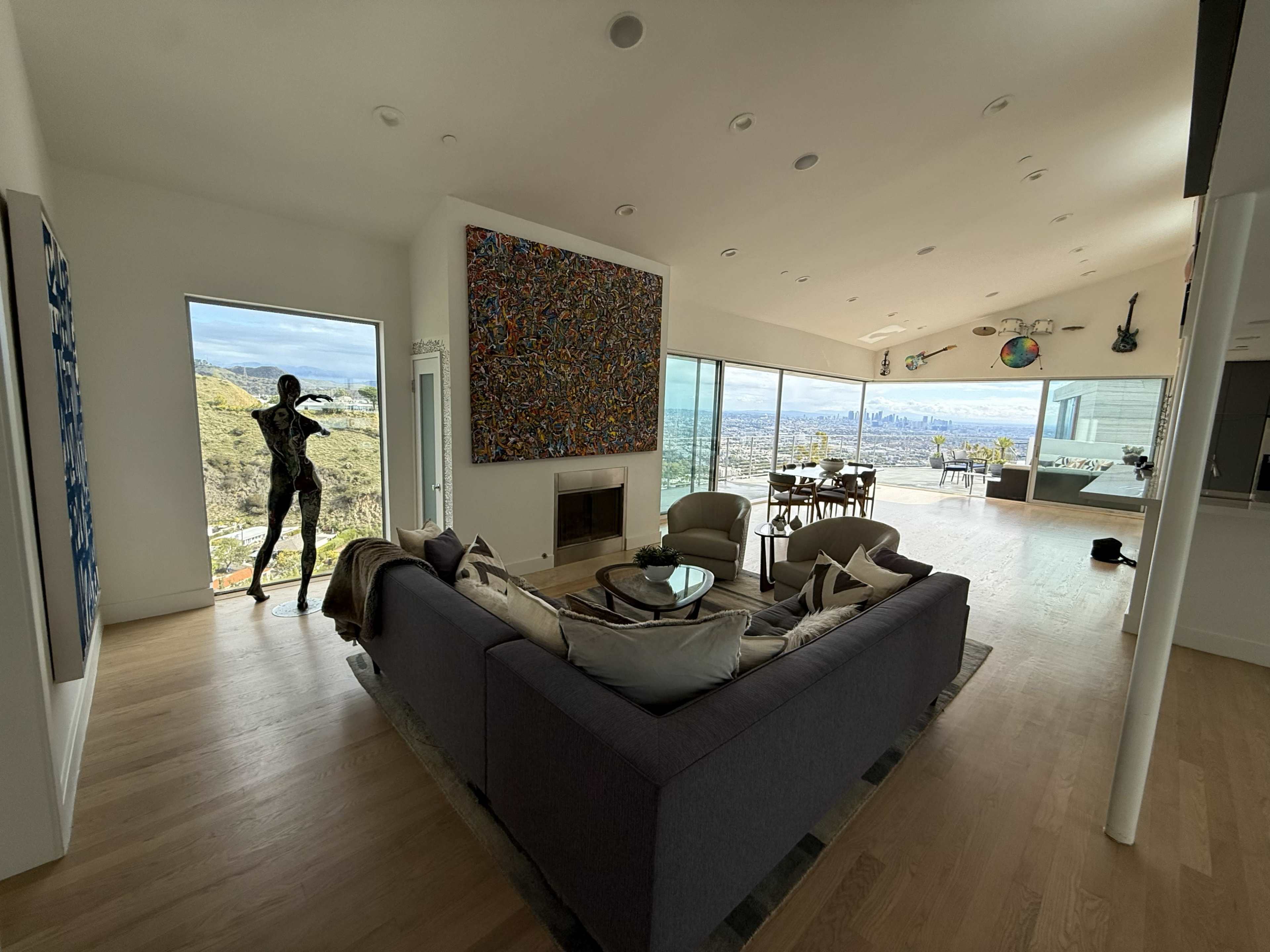 The image shows a modern living room with large windows offering a panoramic view of a city, featuring a sectional sofa, a fireplace, and an abstract painting on the wall.