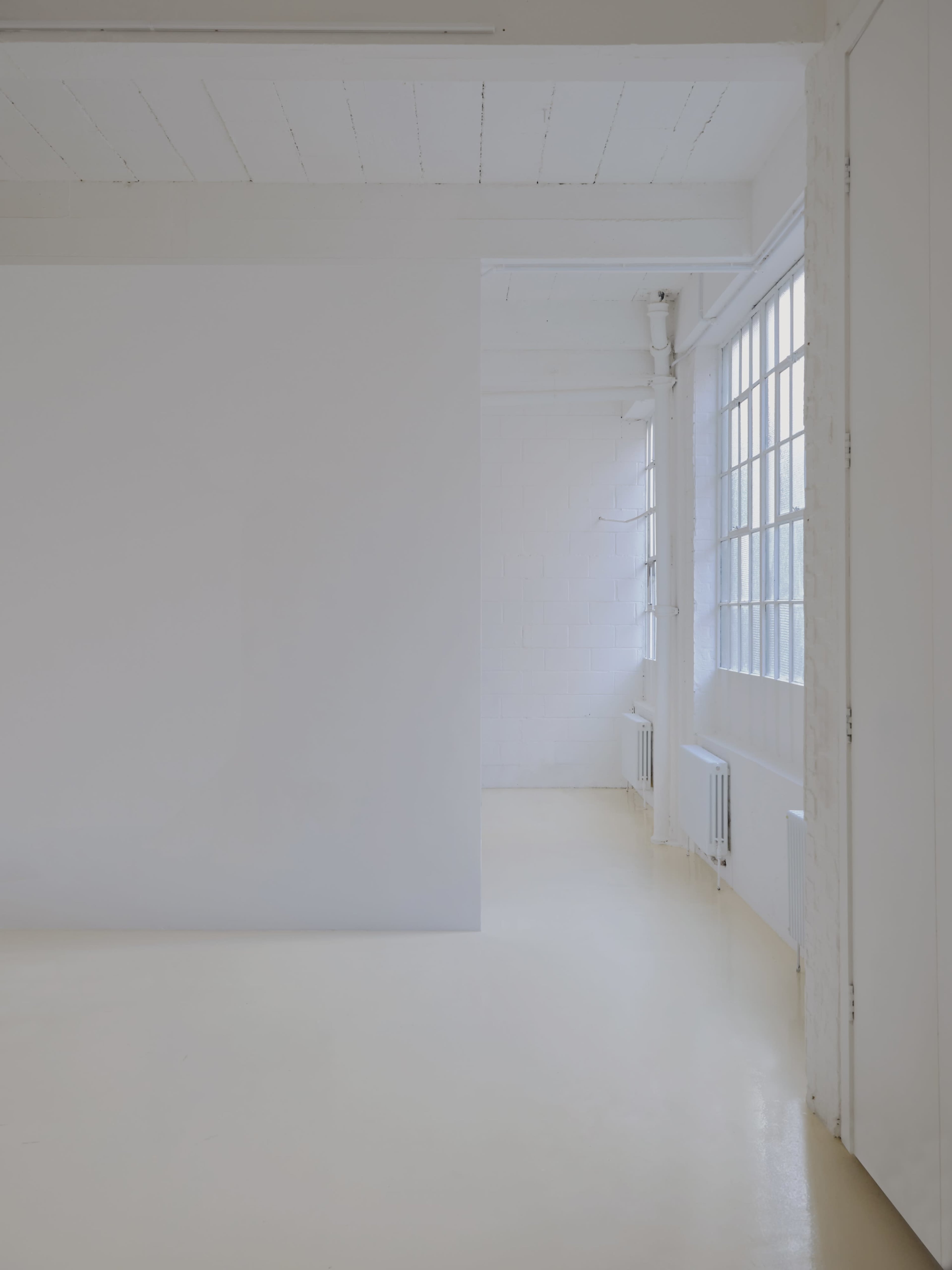 The image shows a bright, minimal interior with white walls, large windows, and a light-colored floor.