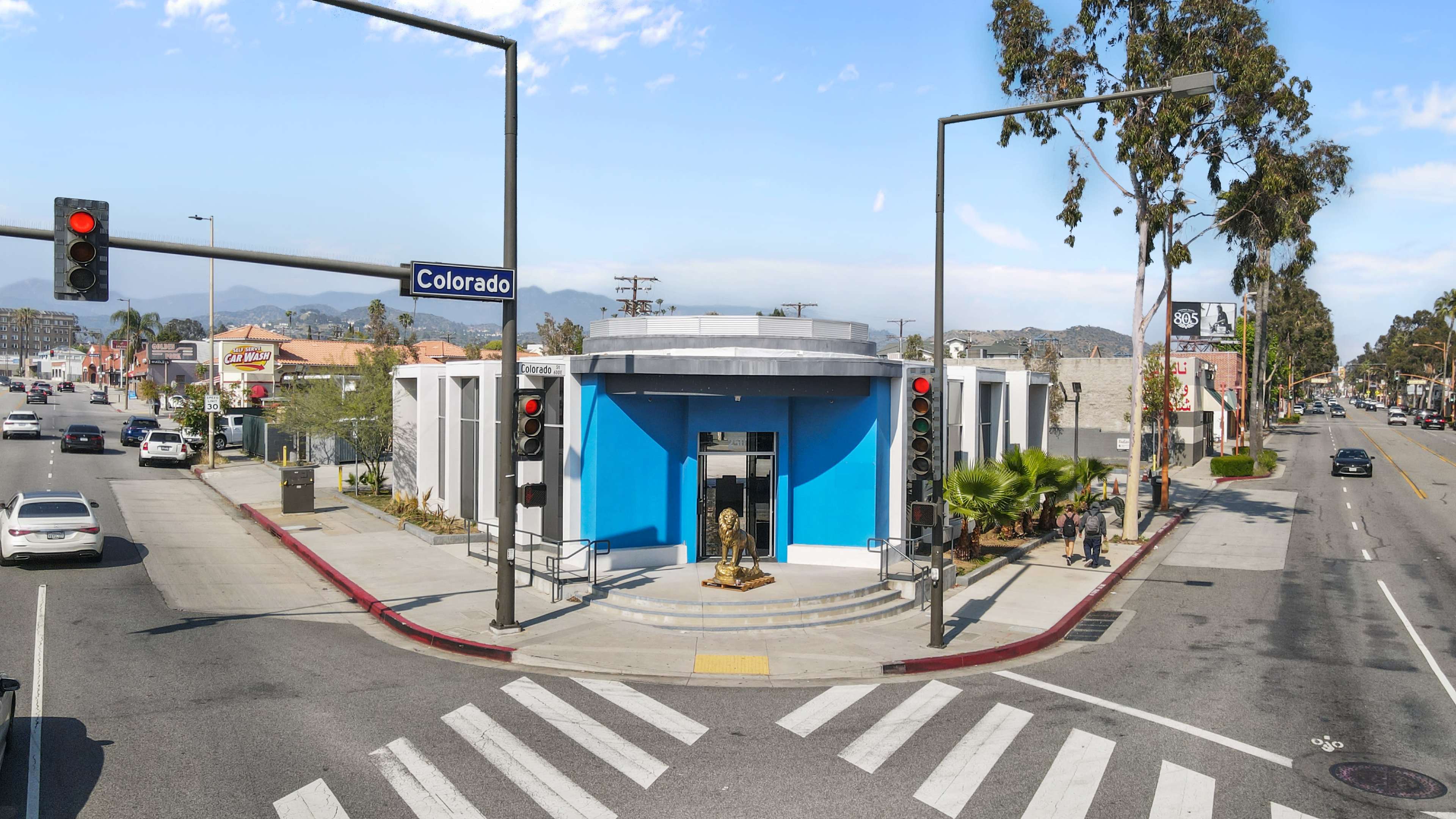 A modern building with a blue facade located at the intersection of Colorado Street and another road, surrounded by palm trees and a few pedestrians.