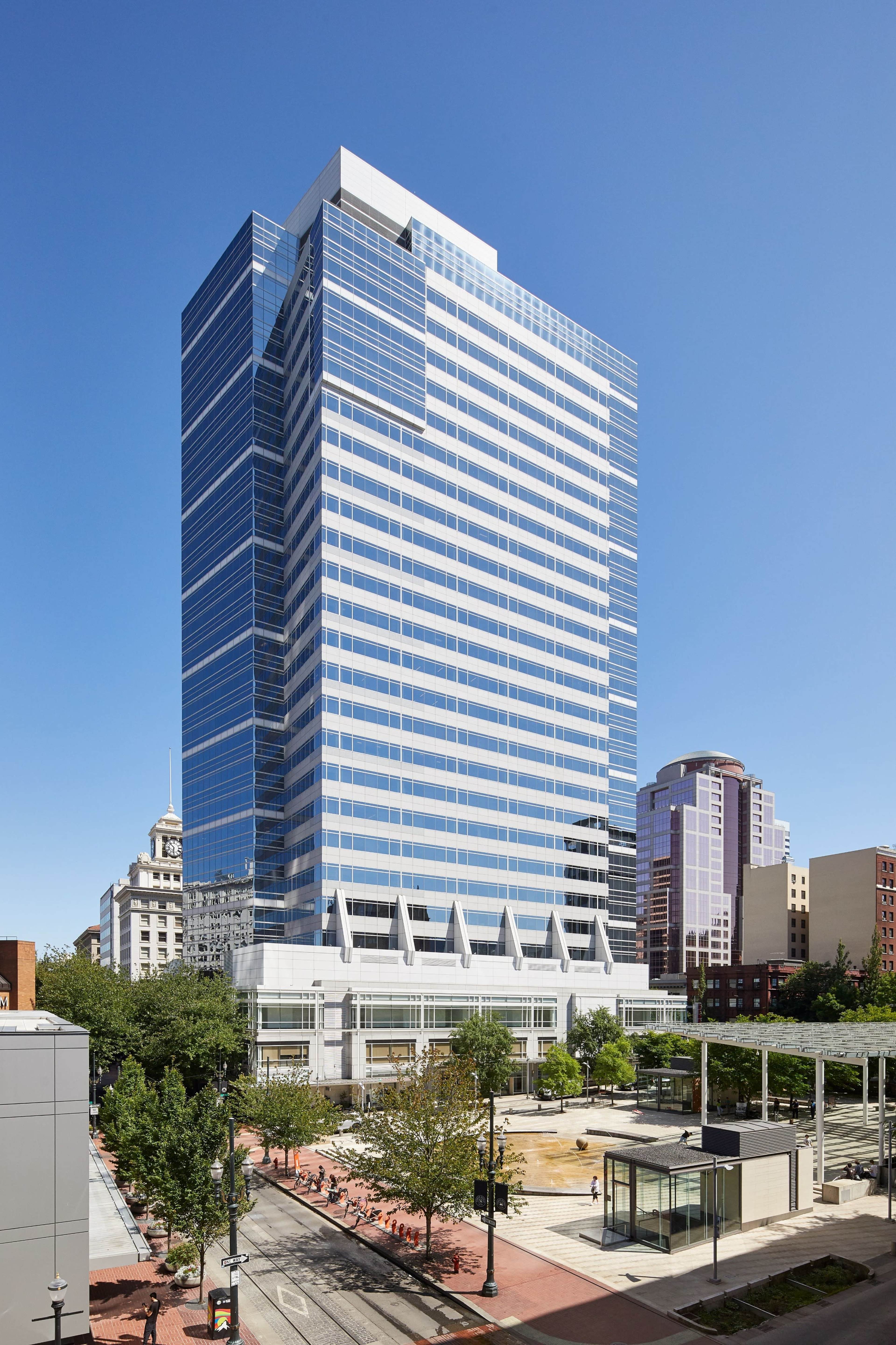 A tall glass office building stands prominently against a clear blue sky, surrounded by trees and a public plaza.