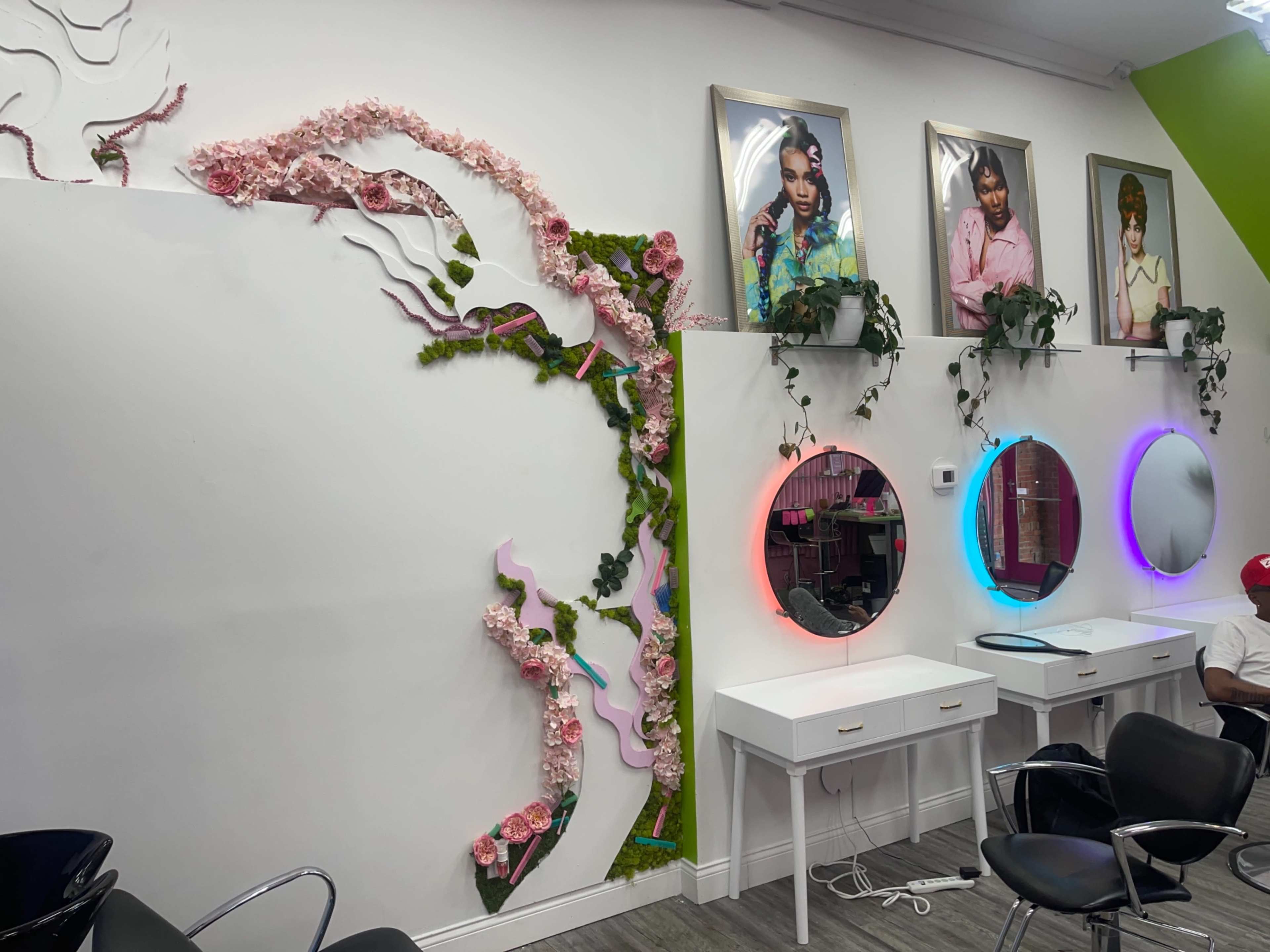 A hair salon interior featuring a wall adorned with floral decorations and framed portraits, alongside two illuminated mirrors and a white vanity.