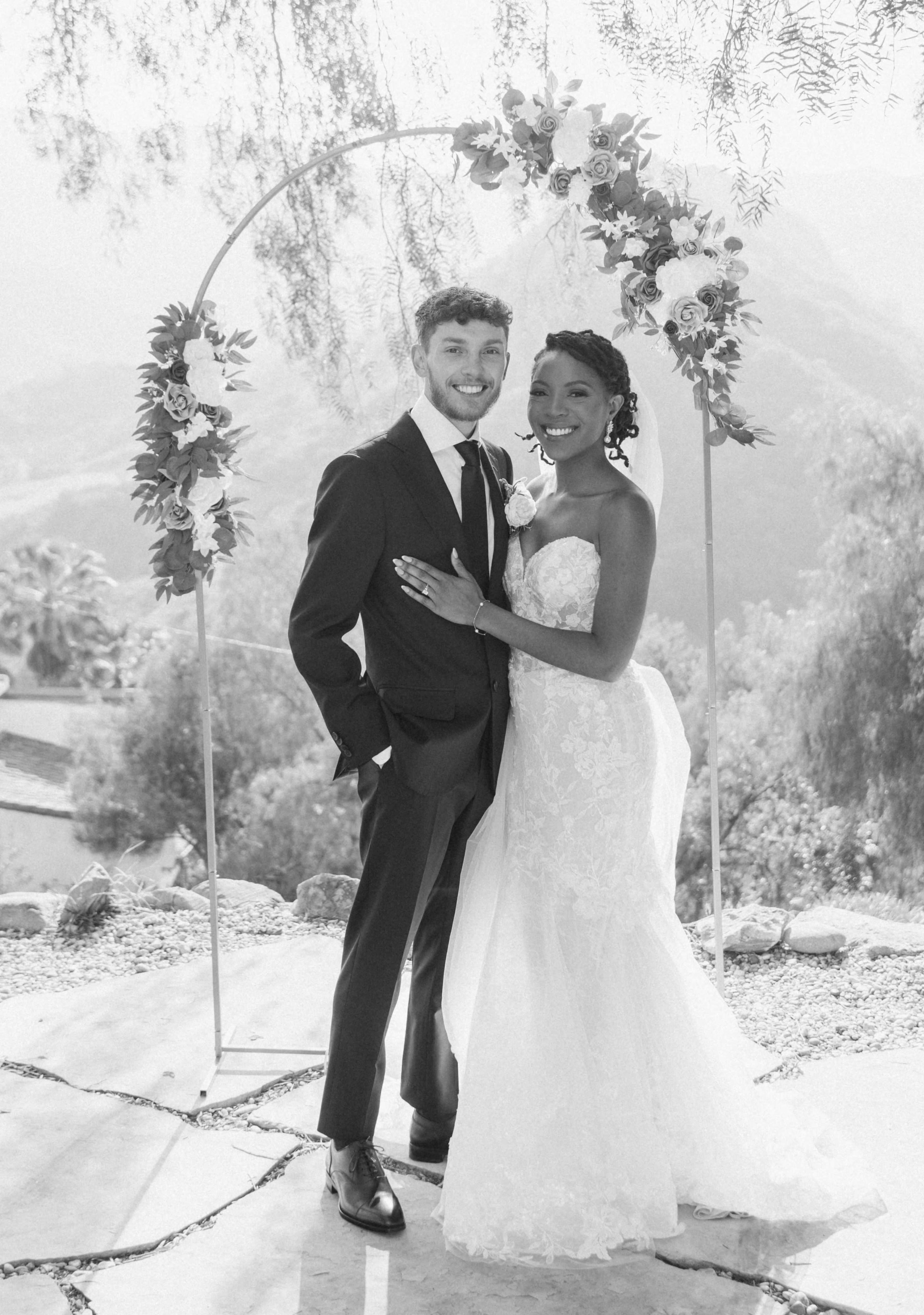 A couple stands together in formal attire under an arch decorated with flowers, against a scenic mountainous backdrop.