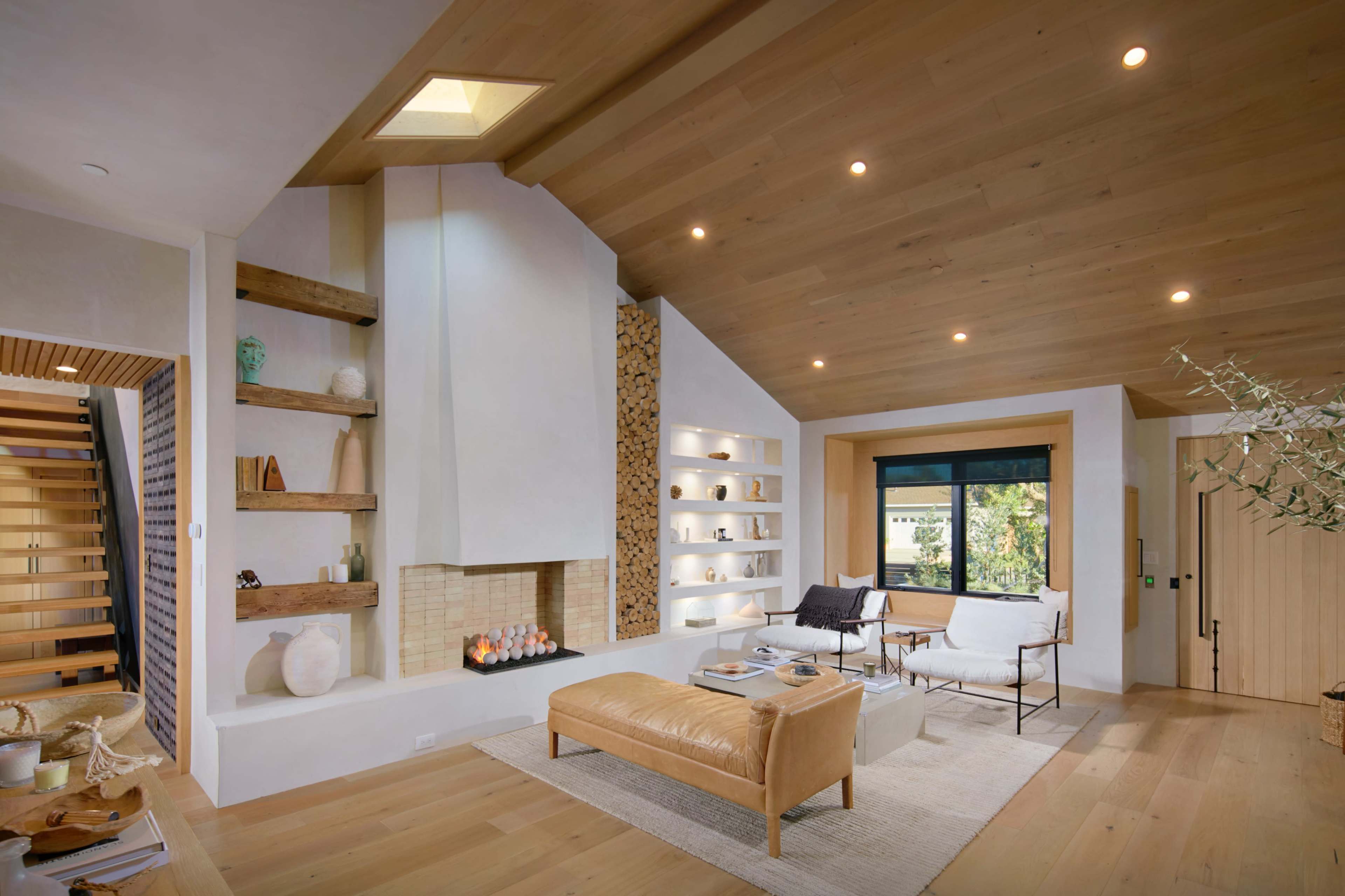 A modern living room featuring a white fireplace, built-in shelves with decorative items, a leather ottoman, and large windows allowing natural light.
