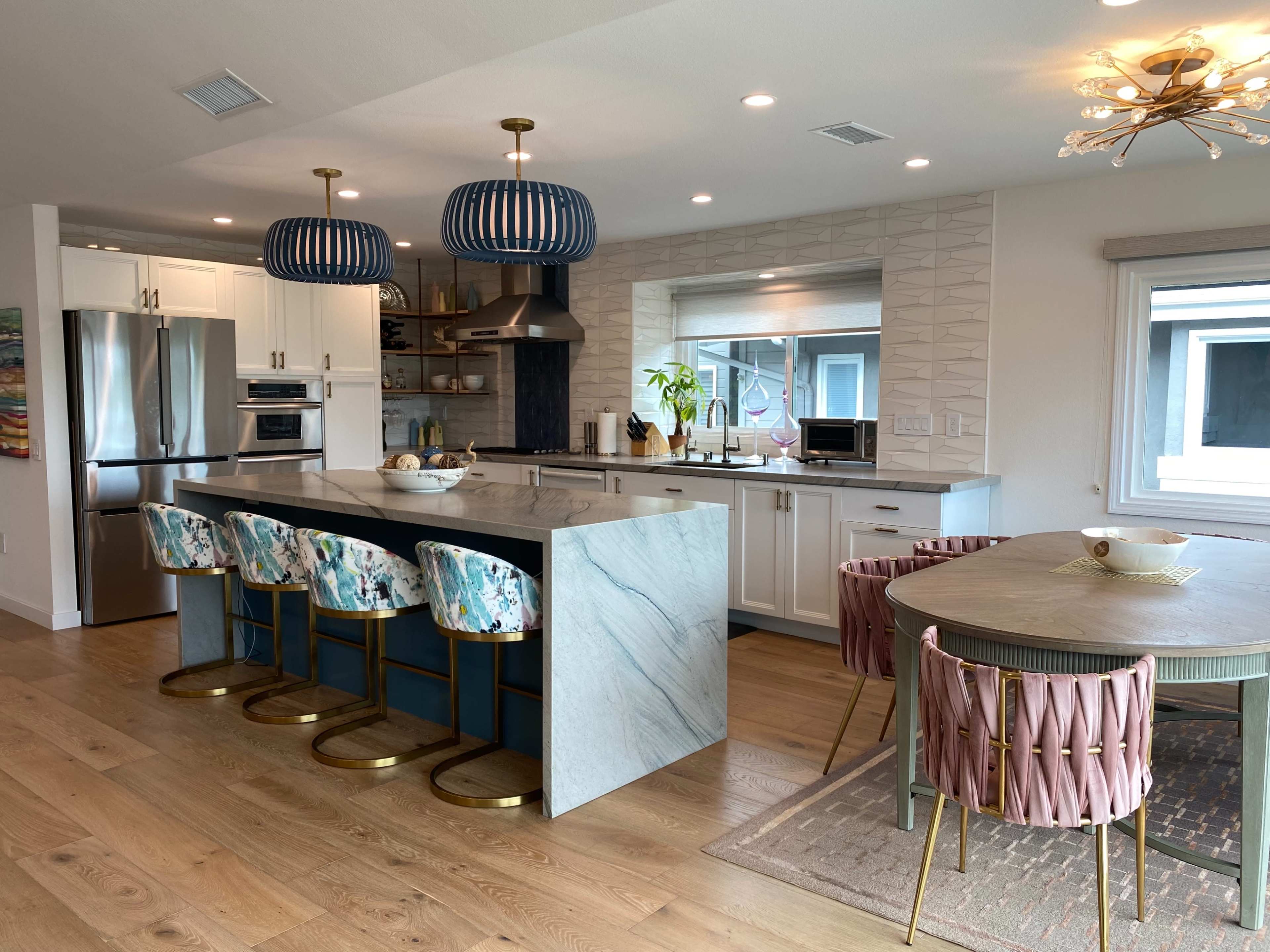 A modern kitchen featuring a marble island with four stools, a dining table with pink and green chairs, and decorative pendant lights.