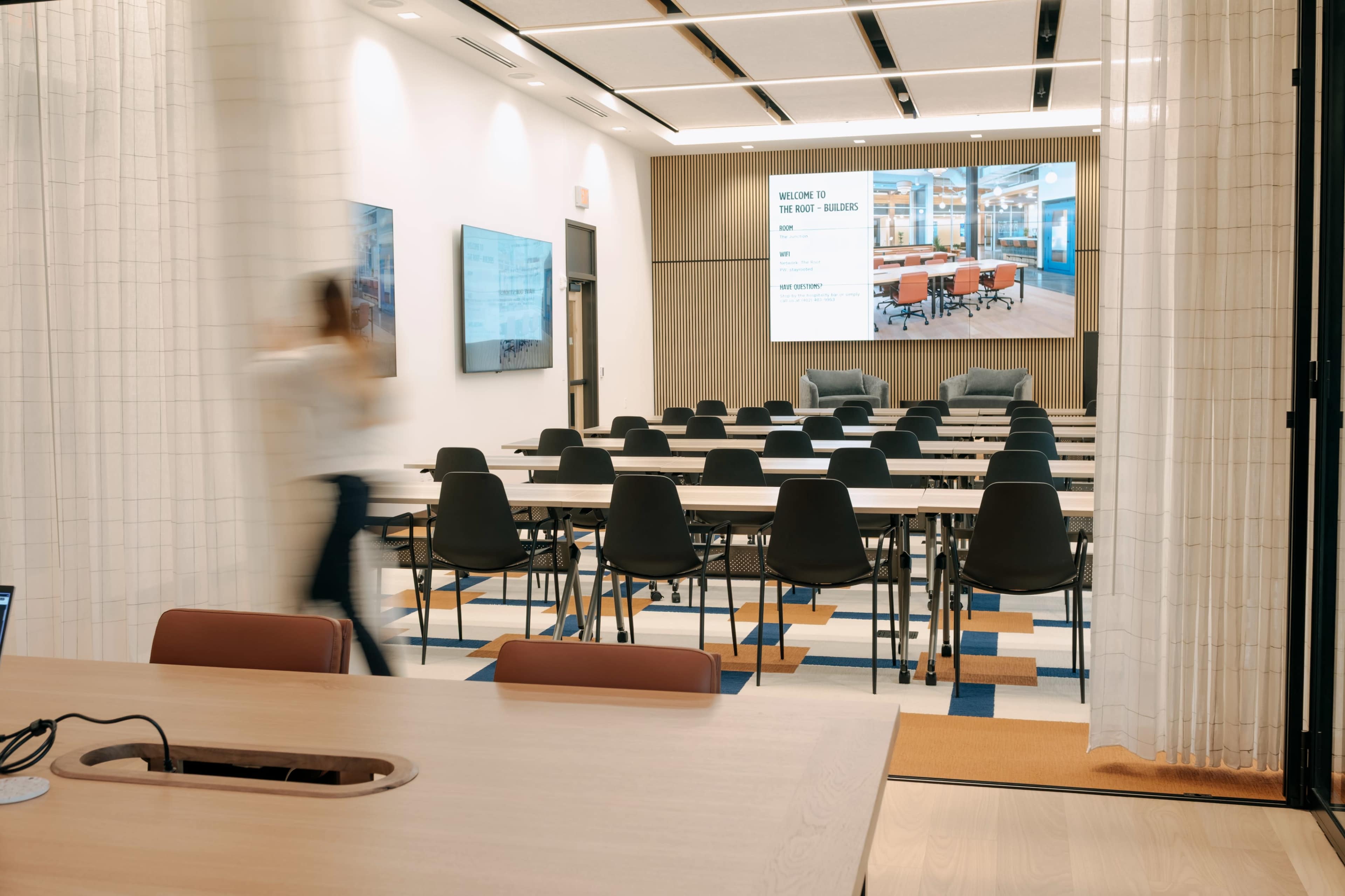 A modern conference room features rows of chairs and tables facing a large screen, with a person walking past in the foreground.