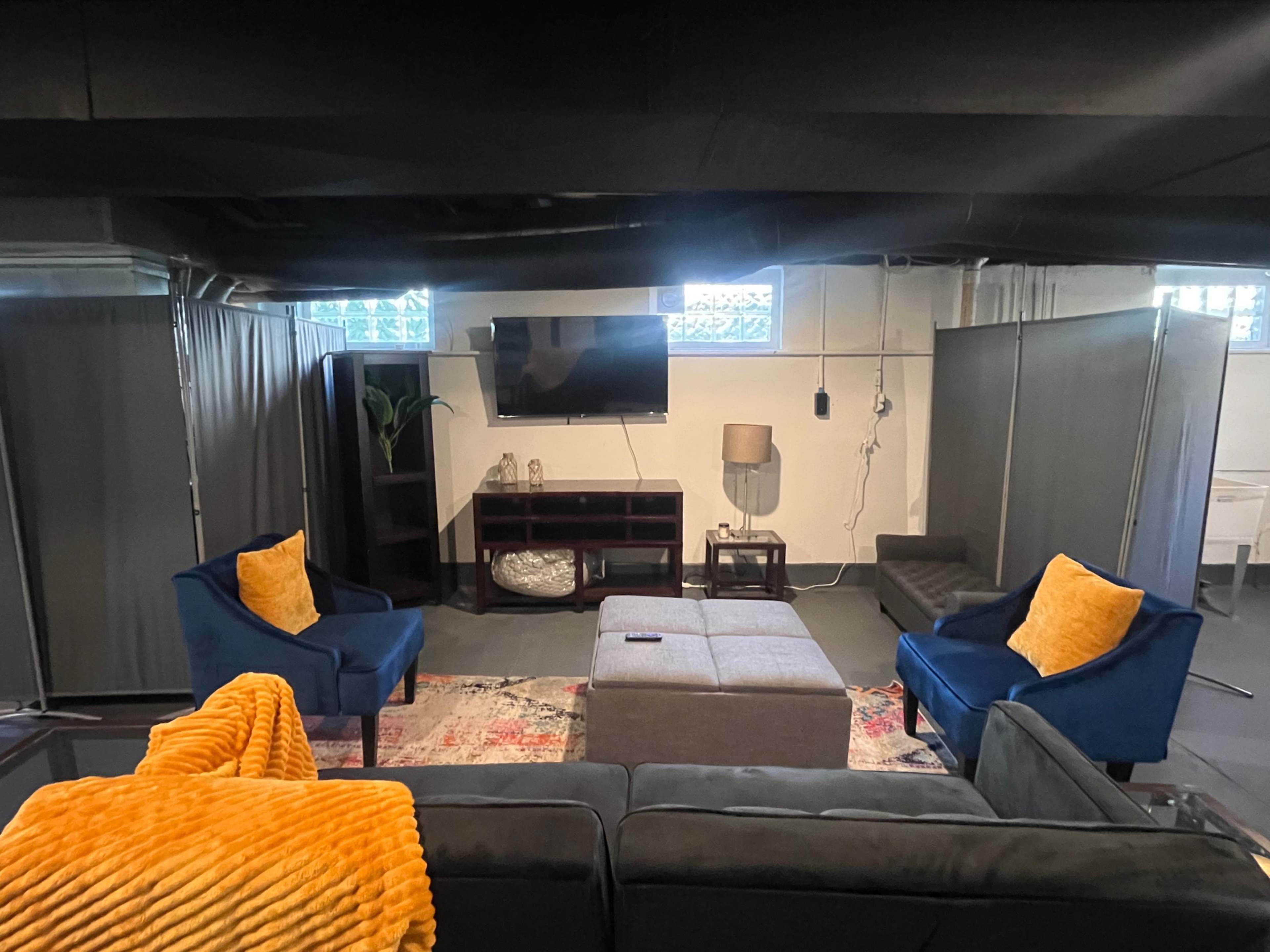 The image shows a finished basement living room with two blue armchairs, a gray sectional couch, a coffee table, a television mounted on the wall, and partition screens creating separate spaces.