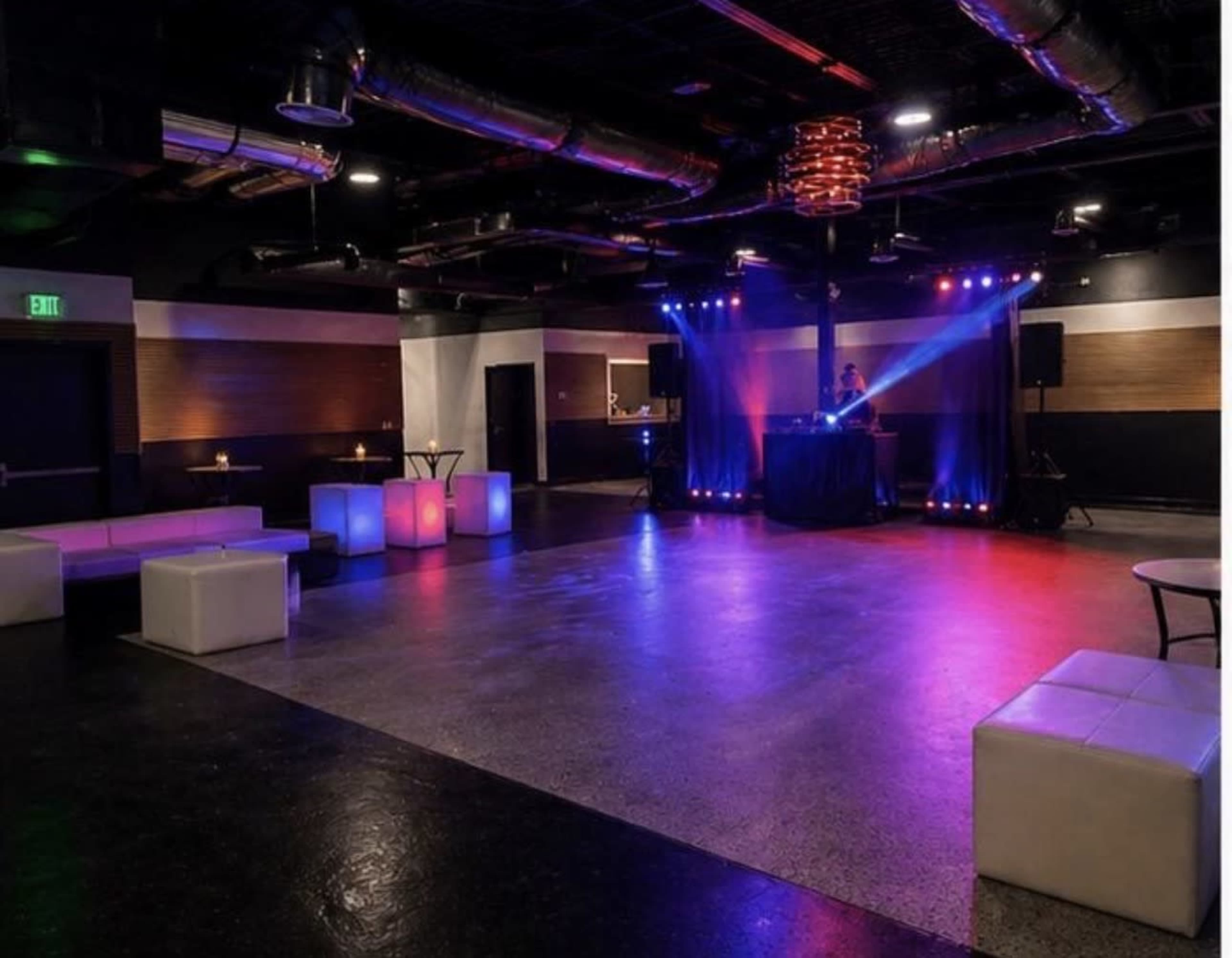 The image shows a dimly lit event space with colorful lighting, a DJ setup, and white seating arrangements.