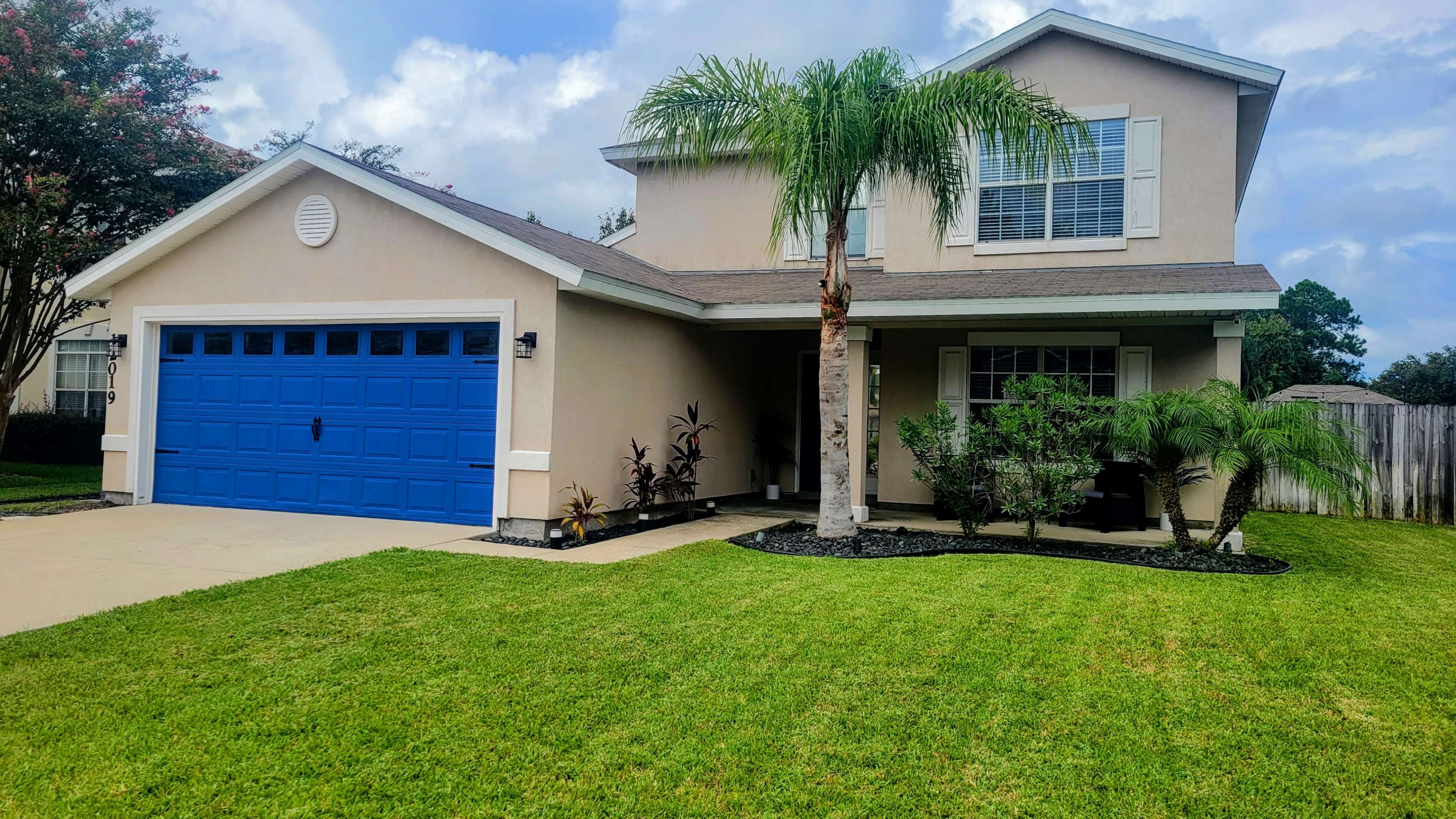 A two-story house with a blue garage door, palm trees, and a well-maintained lawn.