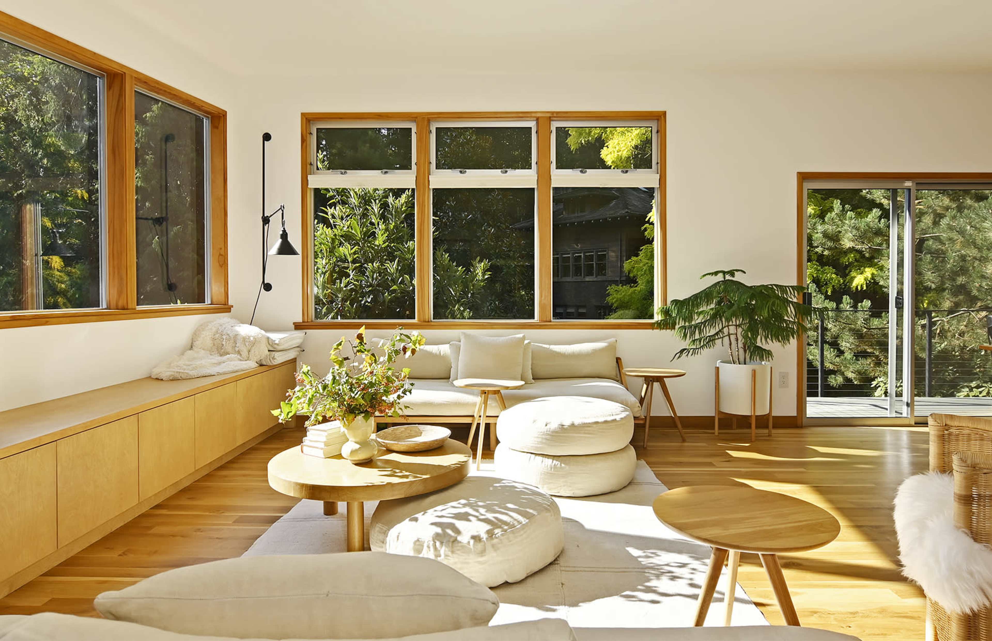 A bright living room features large windows, wooden furniture, and light-colored decor, creating an open and airy space.