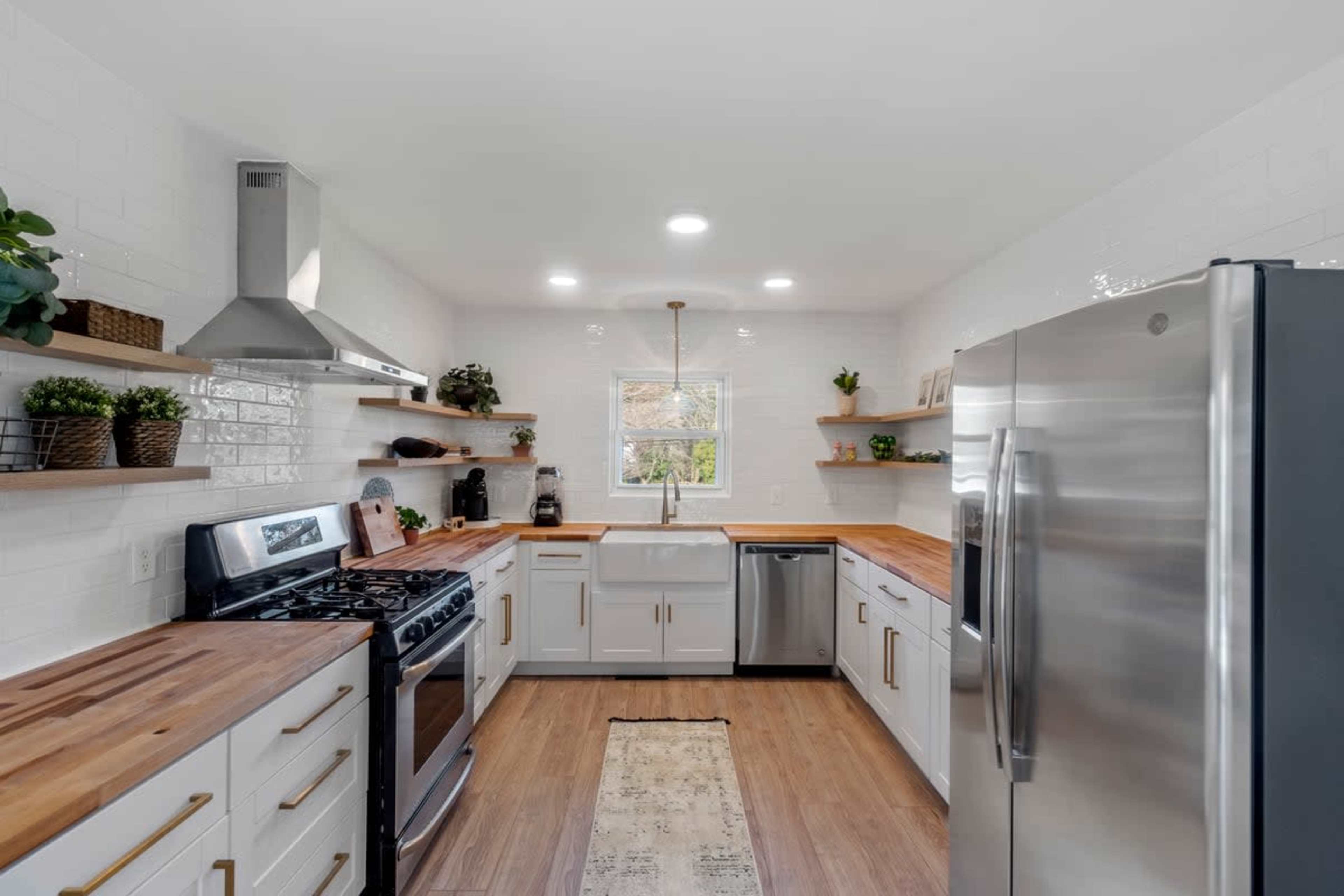 A modern kitchen with stainless steel appliances, white cabinetry, wooden countertops, and open shelving with plants and kitchen items.