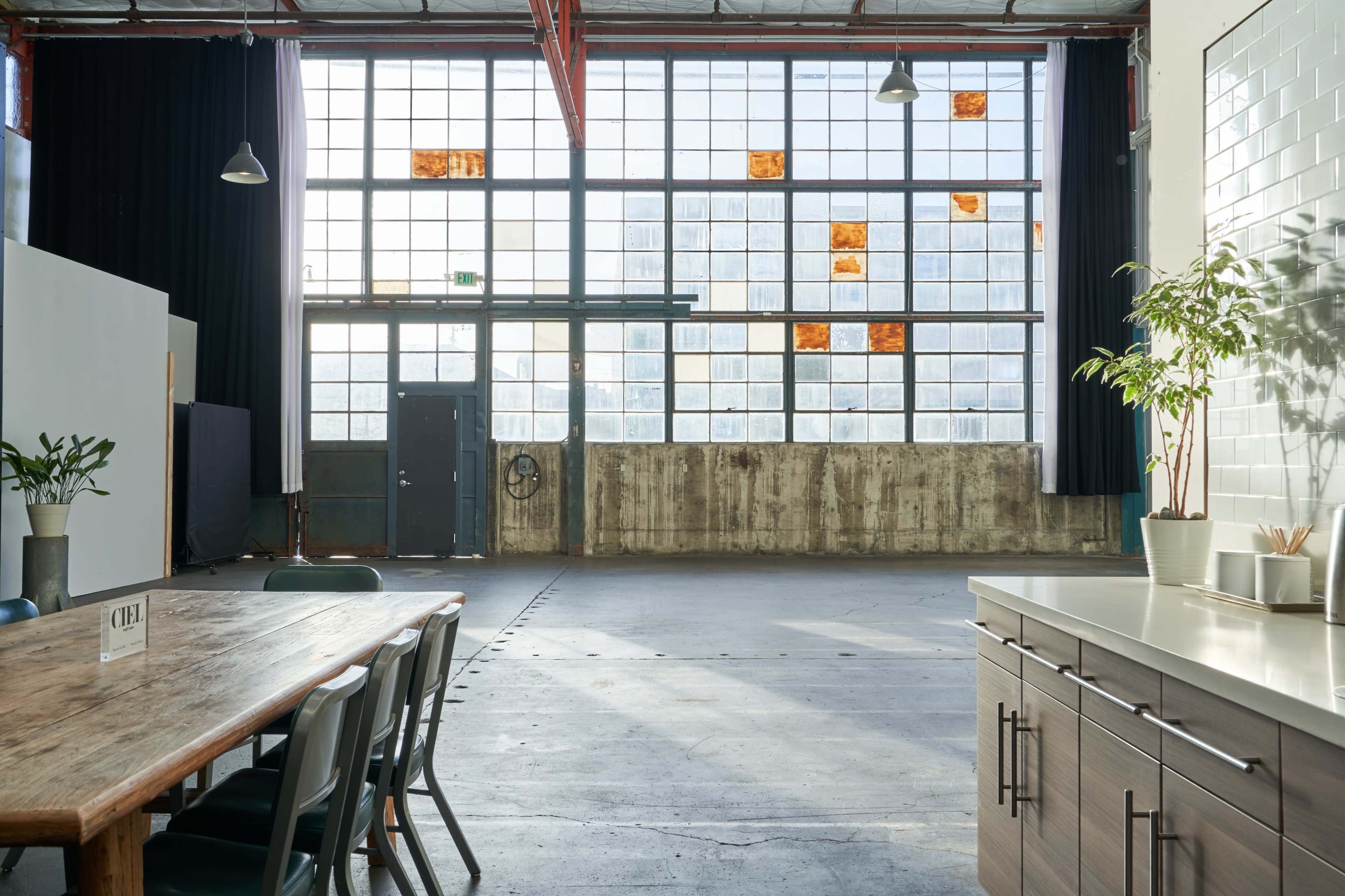 A spacious industrial room with large windows, concrete walls, and a wooden table set with chairs.