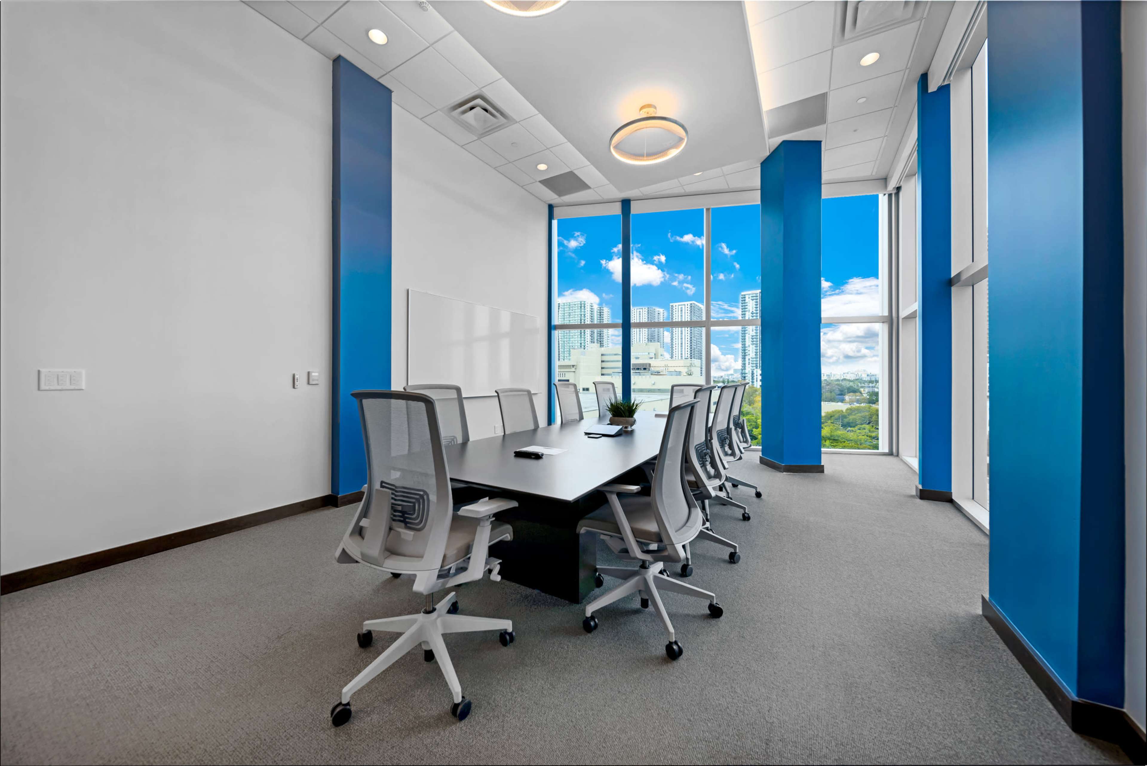A modern conference room features a long table surrounded by ergonomic chairs, with large windows offering a view of a city skyline.