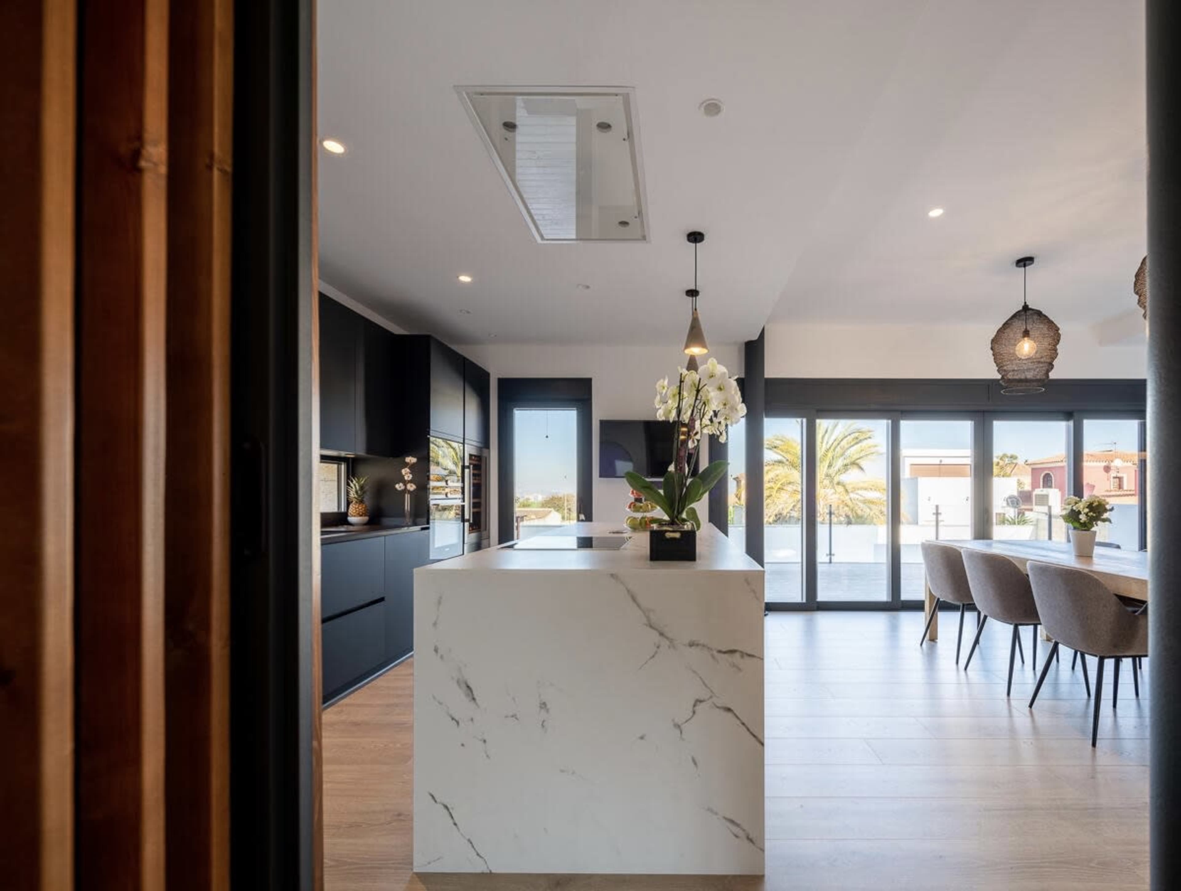 The image shows a modern kitchen with a marble island, black cabinetry, and large windows overlooking a palm tree landscape.