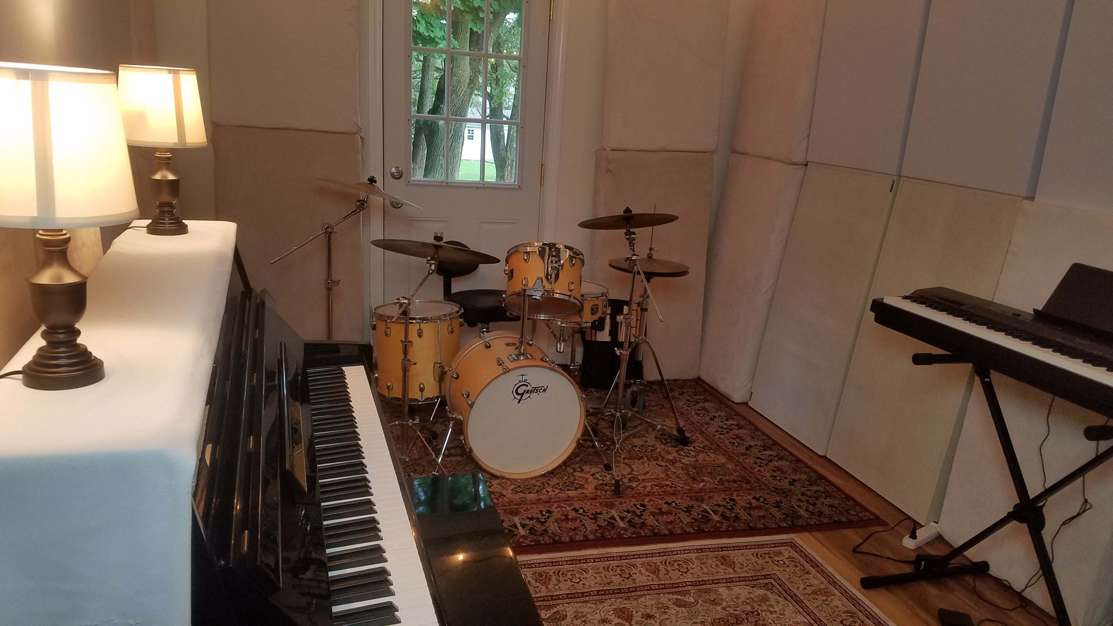 A music room featuring a black piano, a drum set, a keyboard, and lamps, with large windows letting in natural light.