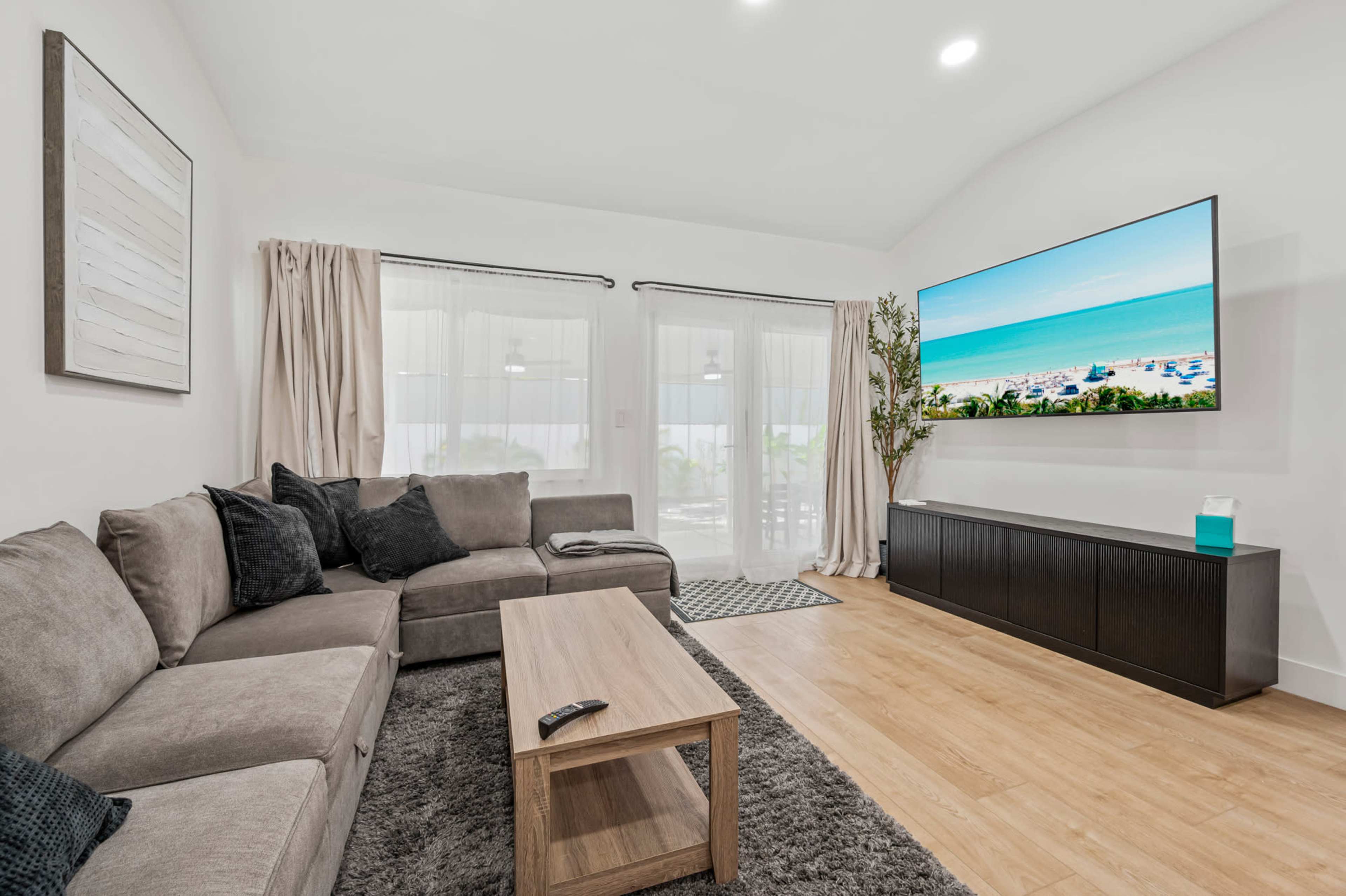 A modern living room features a gray sectional sofa, a wooden coffee table, and a large television mounted on the wall, with light-colored curtains and a rug on the floor.