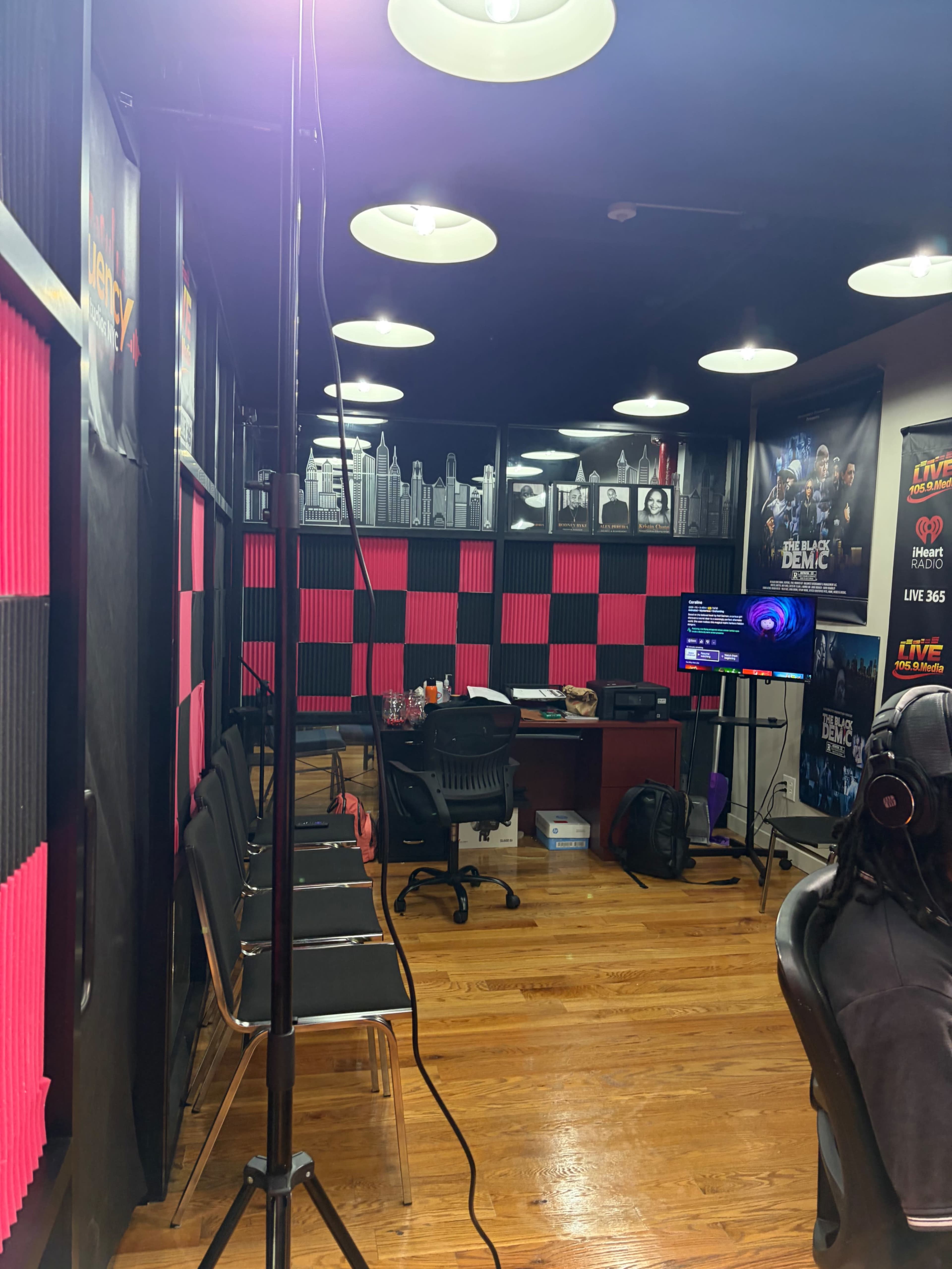 The image shows a radio studio with pink and black soundproofing panels, a desk with equipment, chairs lined up against the wall, and various posters displayed.
