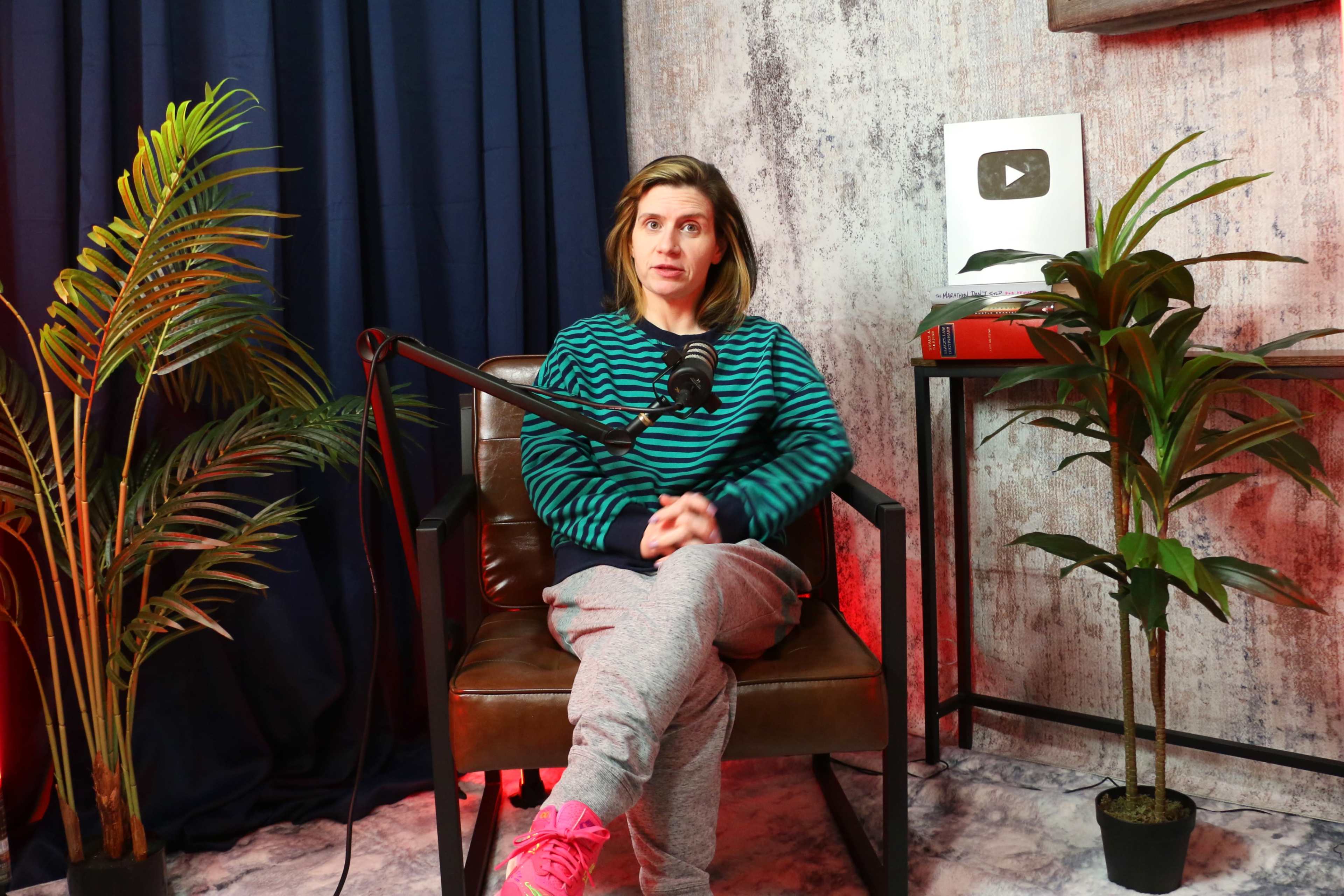 A person sits in a chair with a microphone in front of them, surrounded by potted plants and a backdrop featuring a YouTube play button.