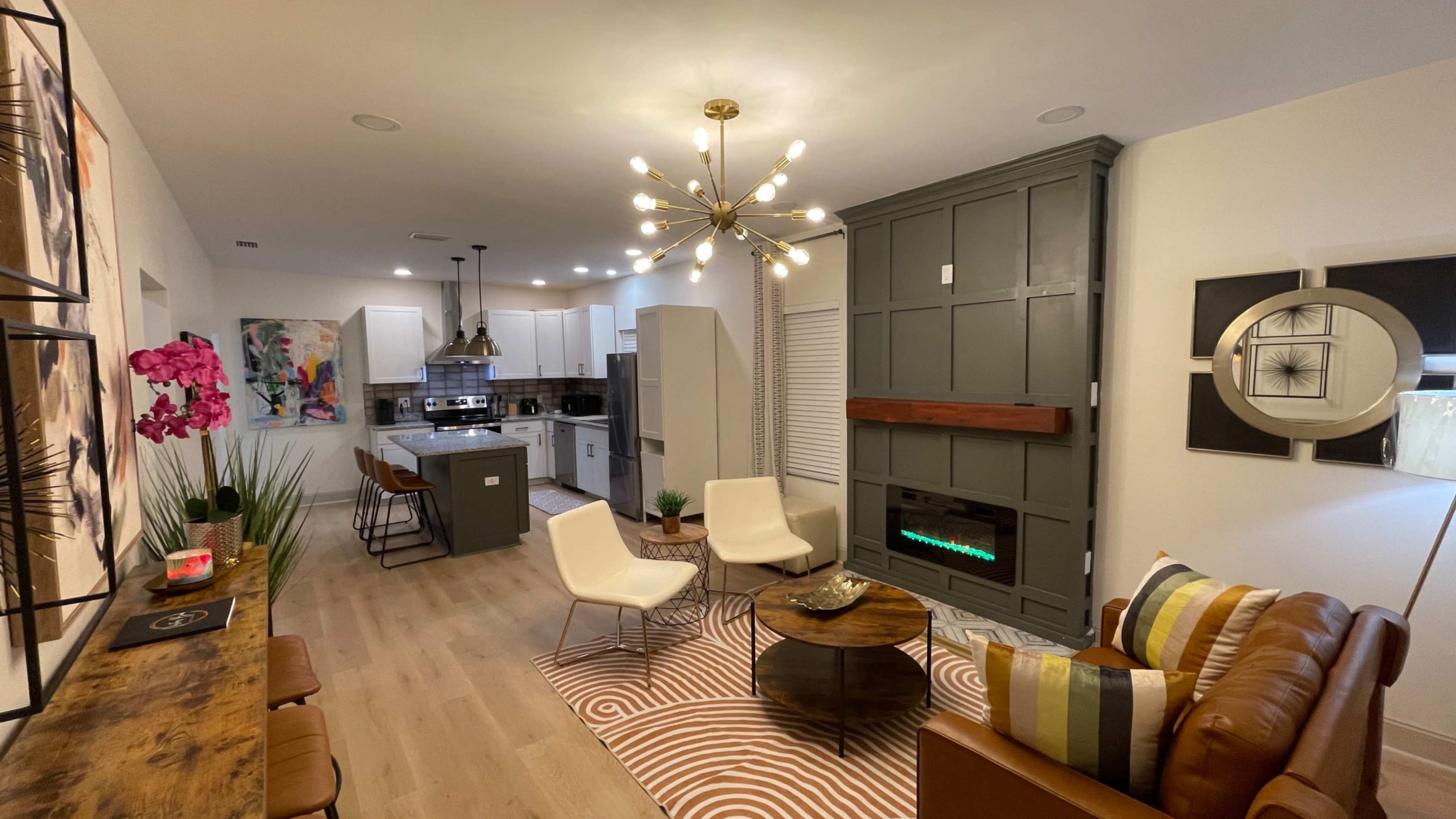 A modern living room with a brown leather sofa, a circular coffee table, and a wall-mounted faux fireplace, connected to an open kitchen with a large island.