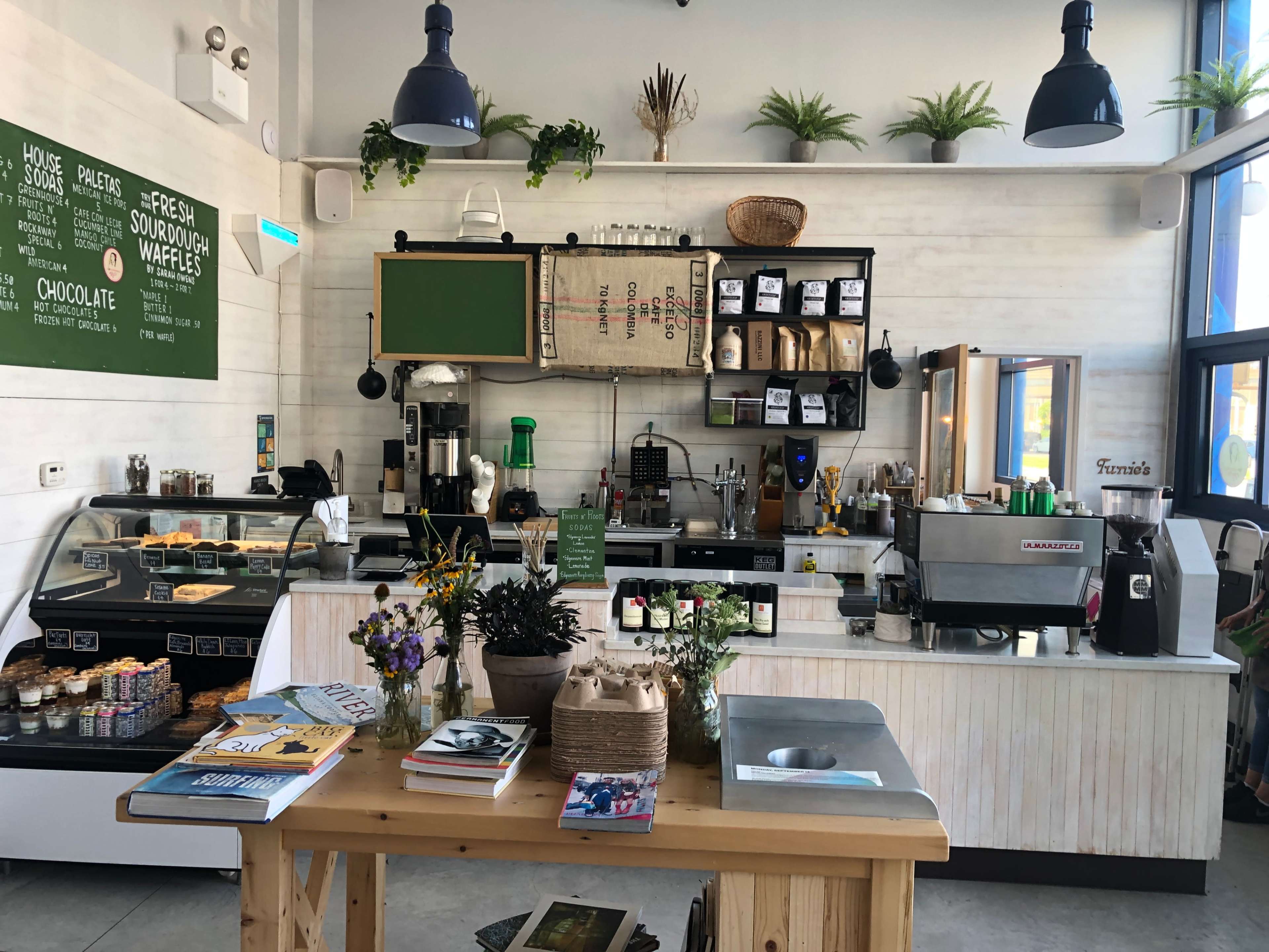 A modern café interior with a display case, coffee equipment, shelves of products, and a wooden table topped with magazines and flowers.