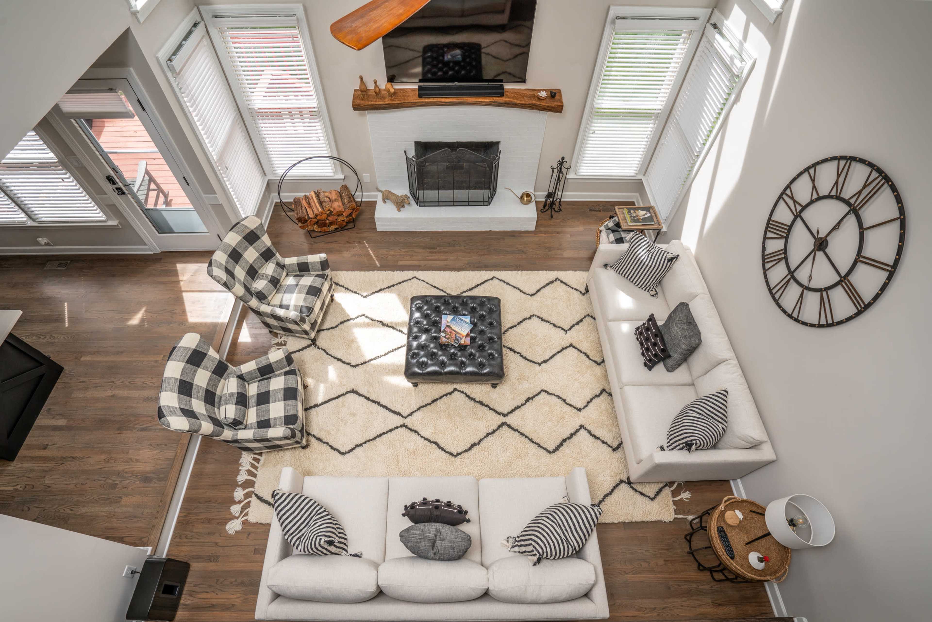 A spacious living room featuring two sofas, two checkered chairs, a coffee table on a patterned rug, and a fireplace with large windows allowing natural light.
