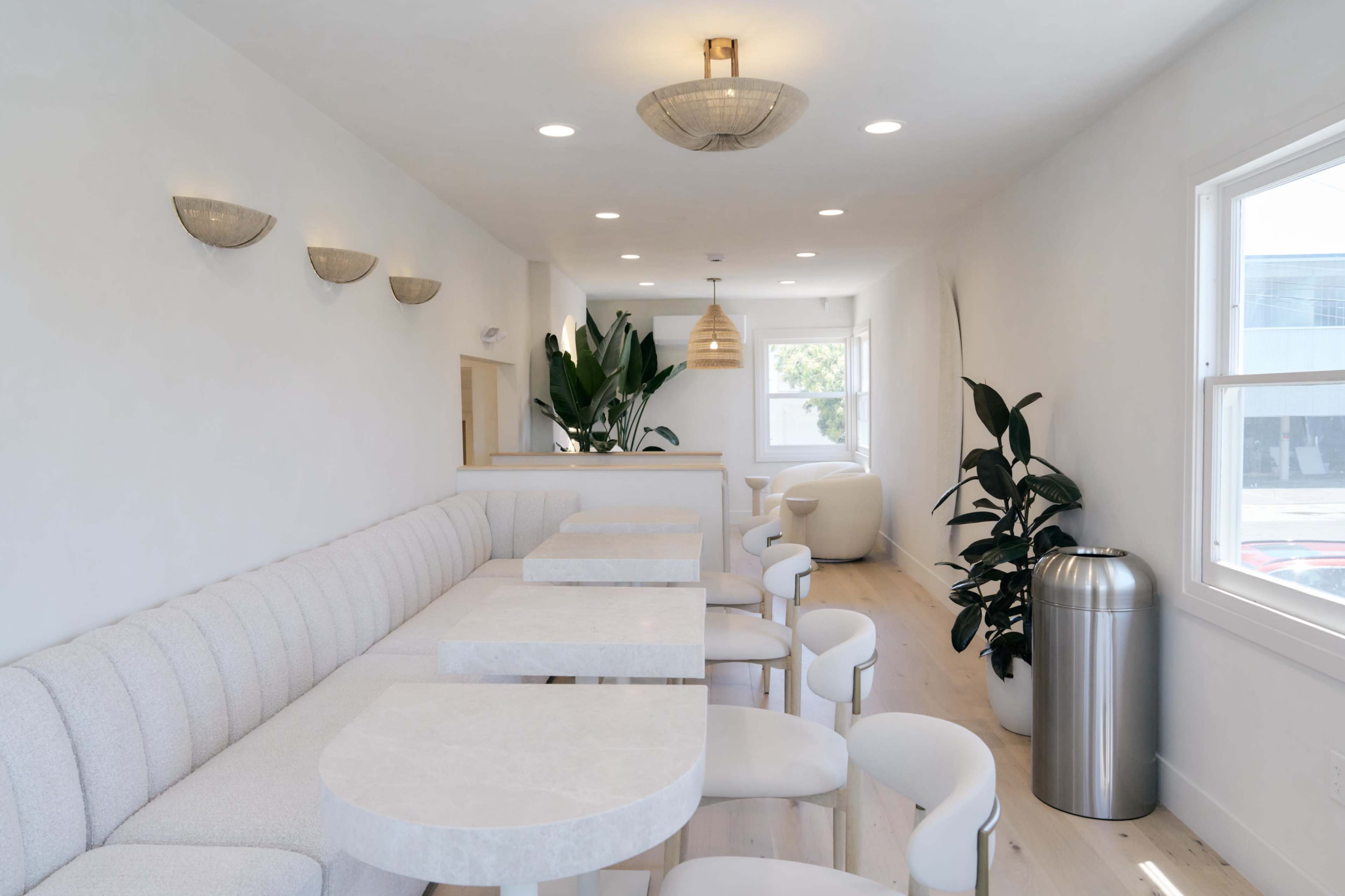 The image shows a modern, minimalistic interior of a café featuring light-colored walls, a cozy seating area with rounded tables, and decorative plants.