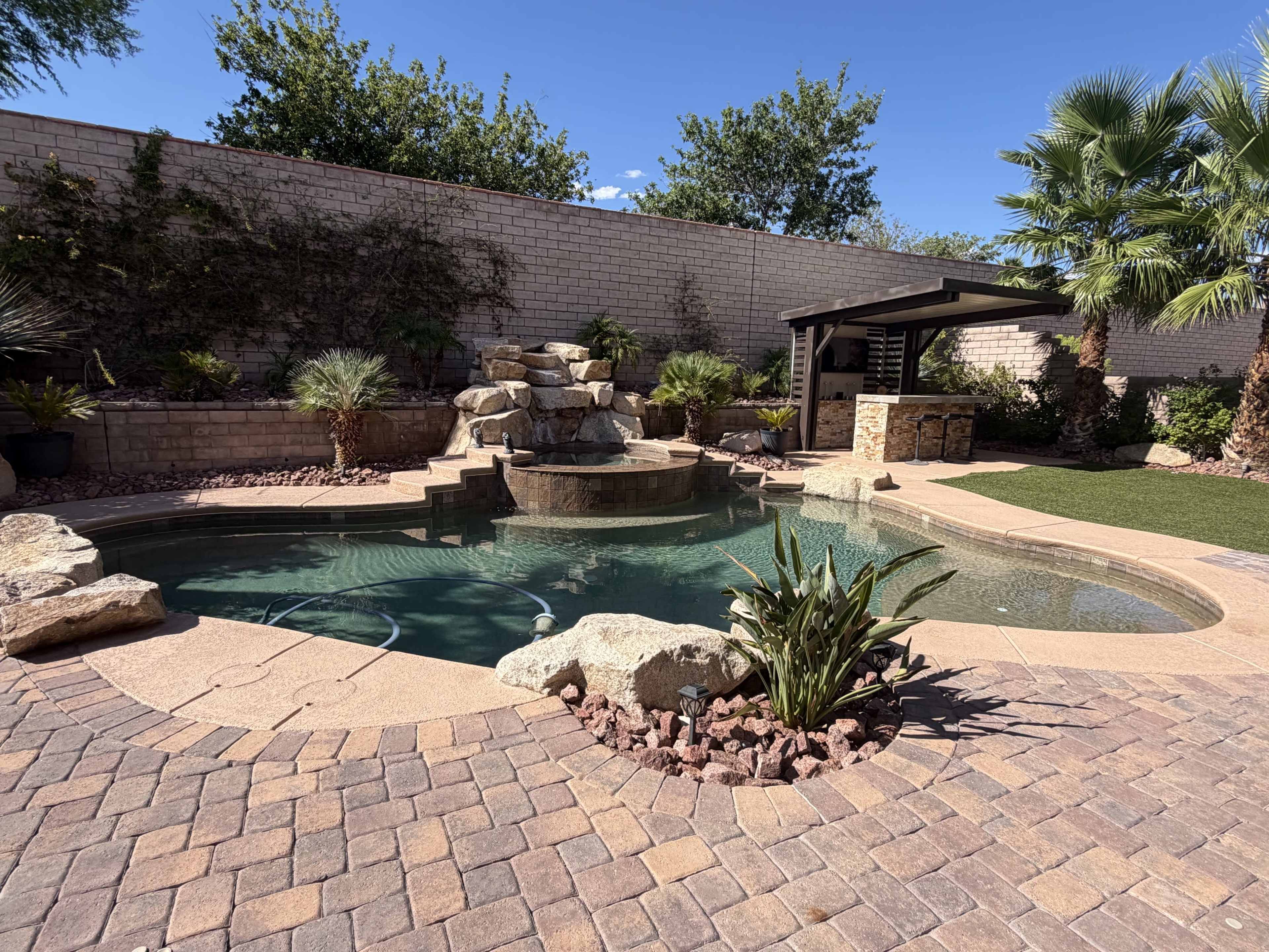 The image shows a landscaped backyard featuring a swimming pool with a rock waterfall, surrounded by palm trees and stone pavers.