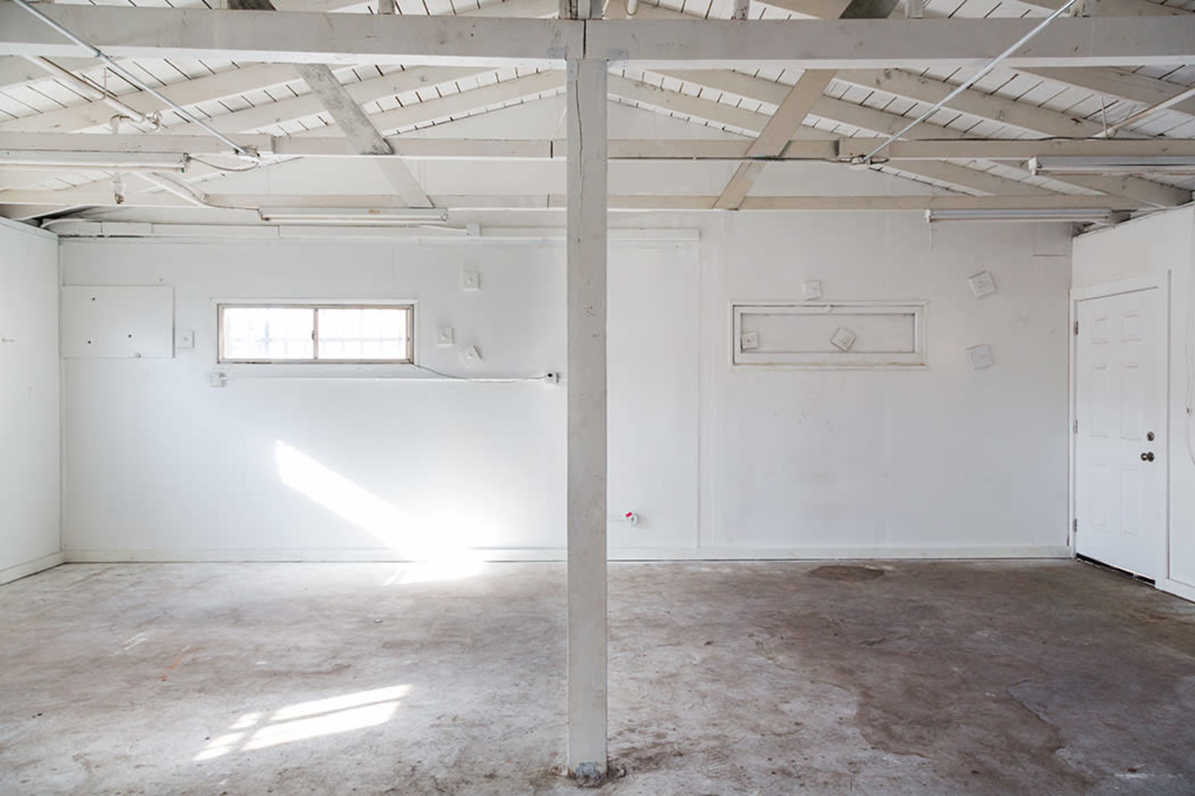 The image shows an empty, well-lit room with white walls, a wooden ceiling, and a concrete floor, featuring a single window and a door.