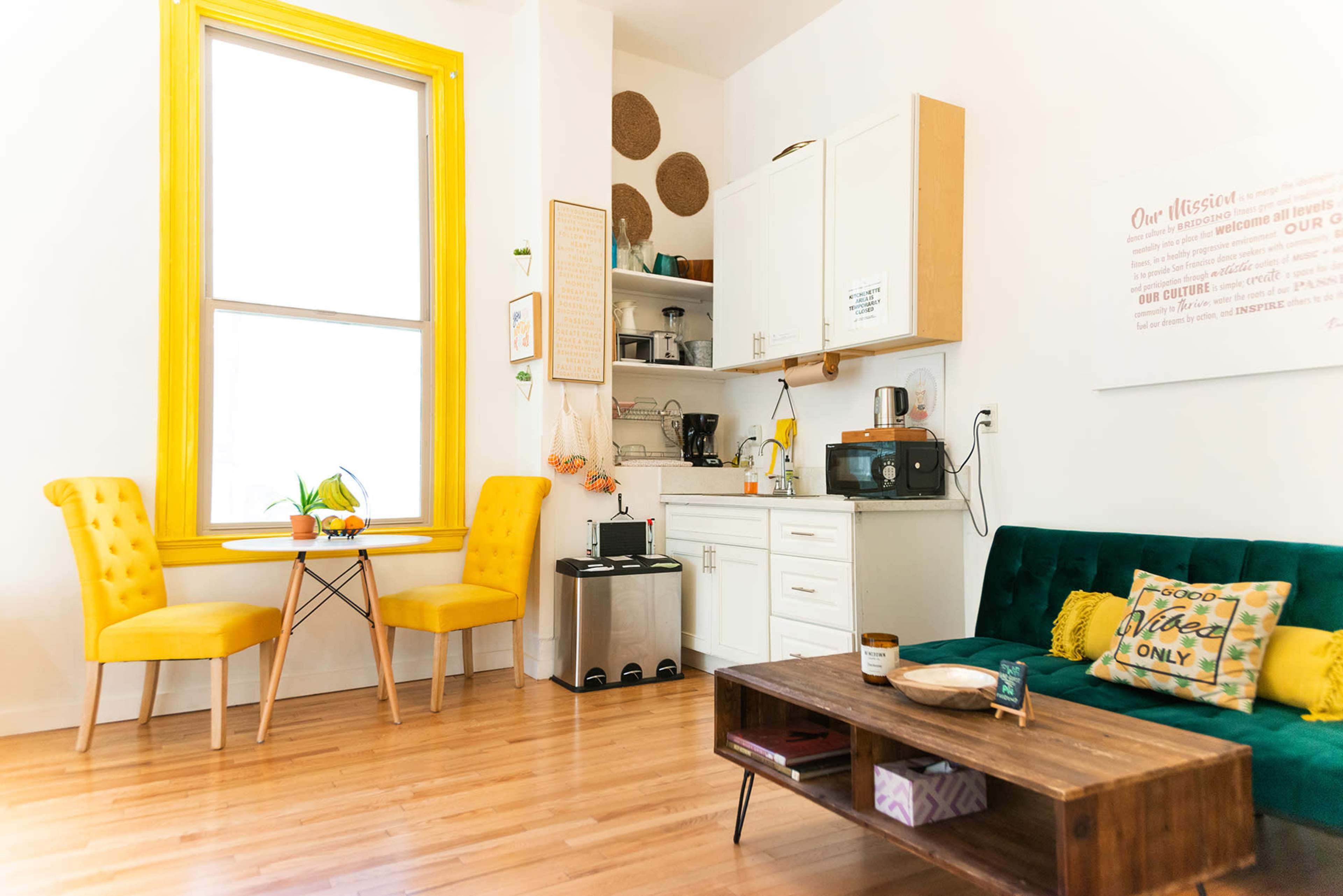 A brightly lit room features a green sofa, a wooden coffee table, yellow chairs at a small dining table, and a compact kitchen area in the corner.