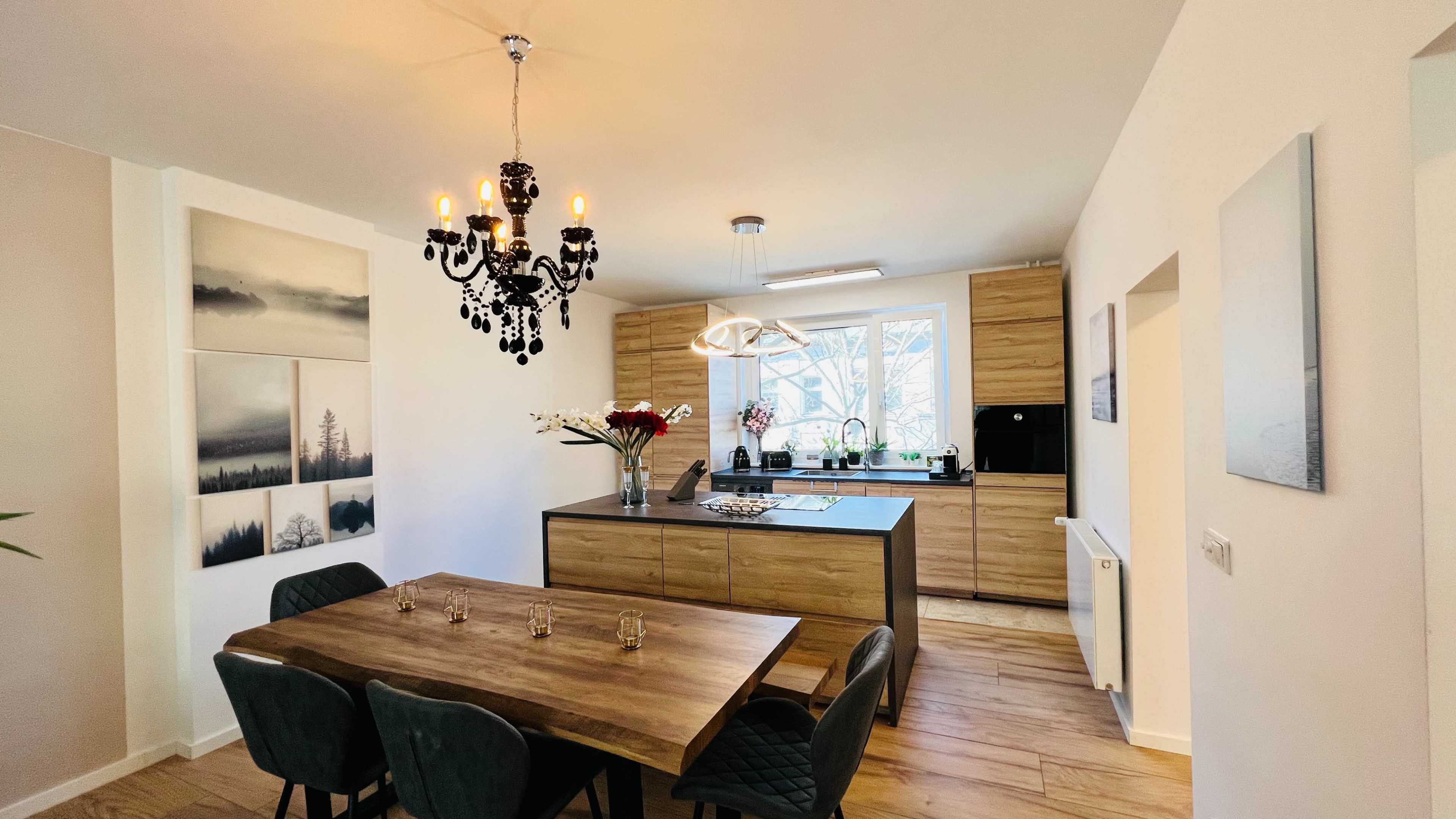 A modern kitchen and dining area with a wooden table, elegant chandelier, and a large window overlooking greenery.