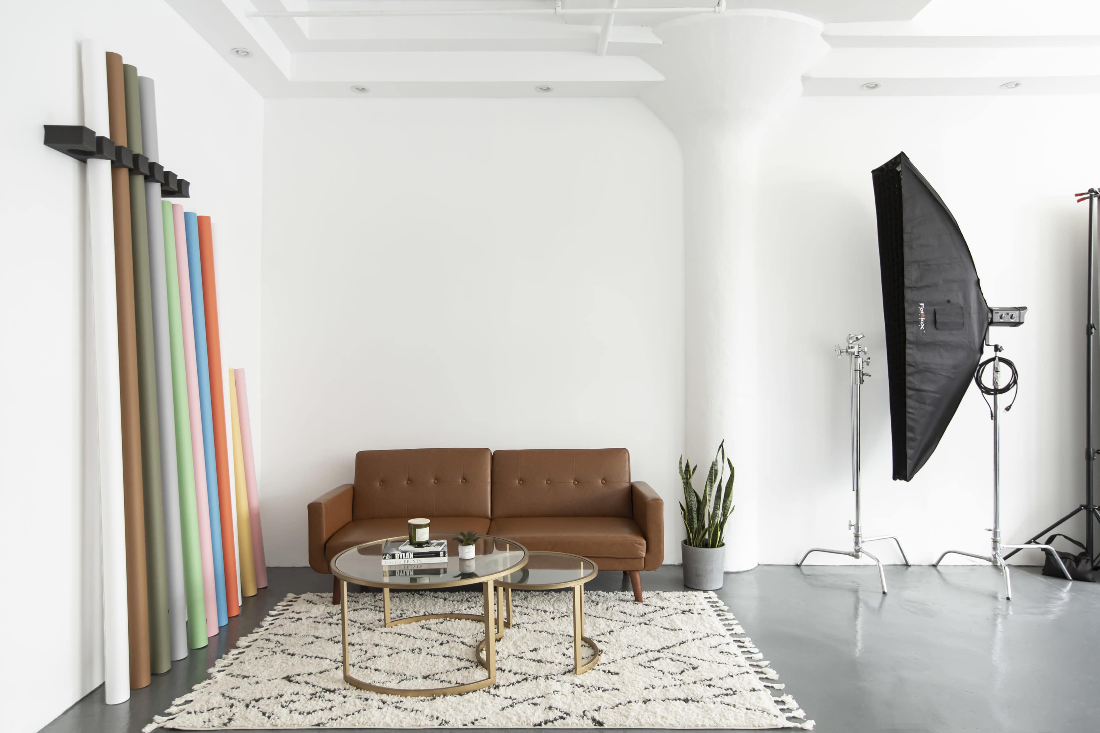 The image shows a modern studio living space with a brown couch, two circular tables, a plant, and a backdrop of colored paper rolls against a white wall.