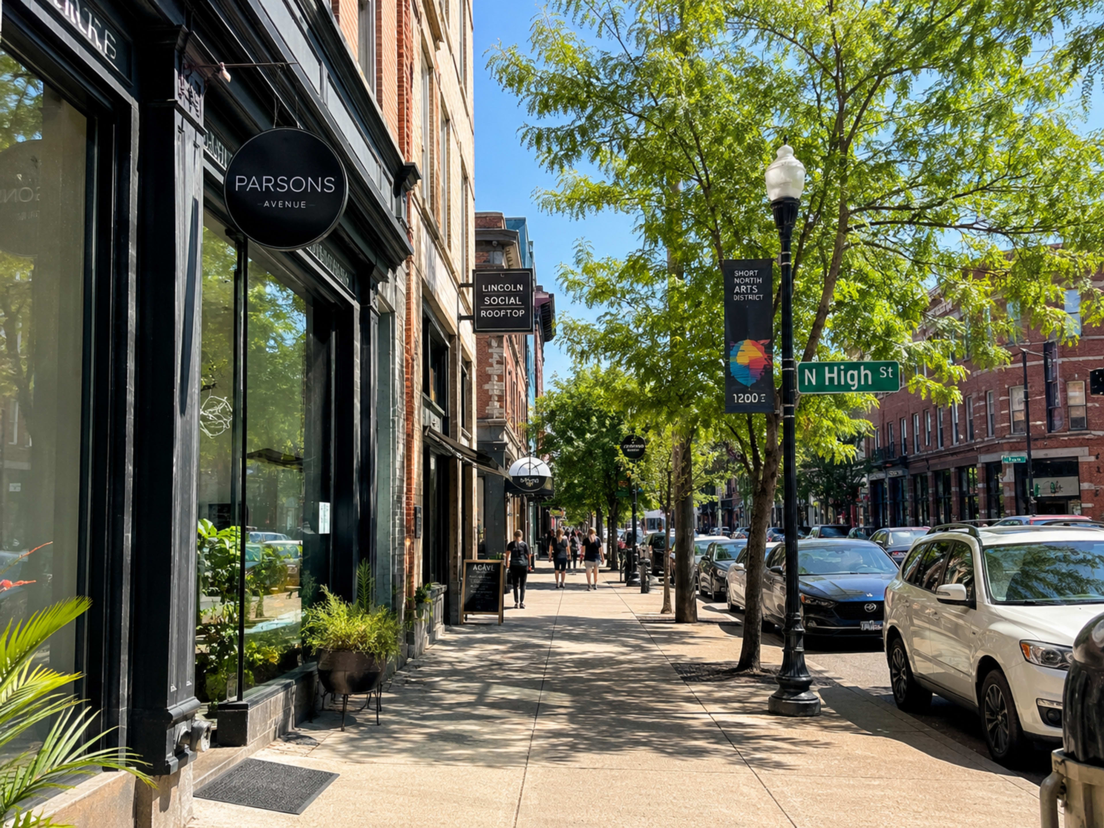 A sunny street scene features storefronts along a sidewalk lined with trees and parked cars, with a street sign indicating North High Street.