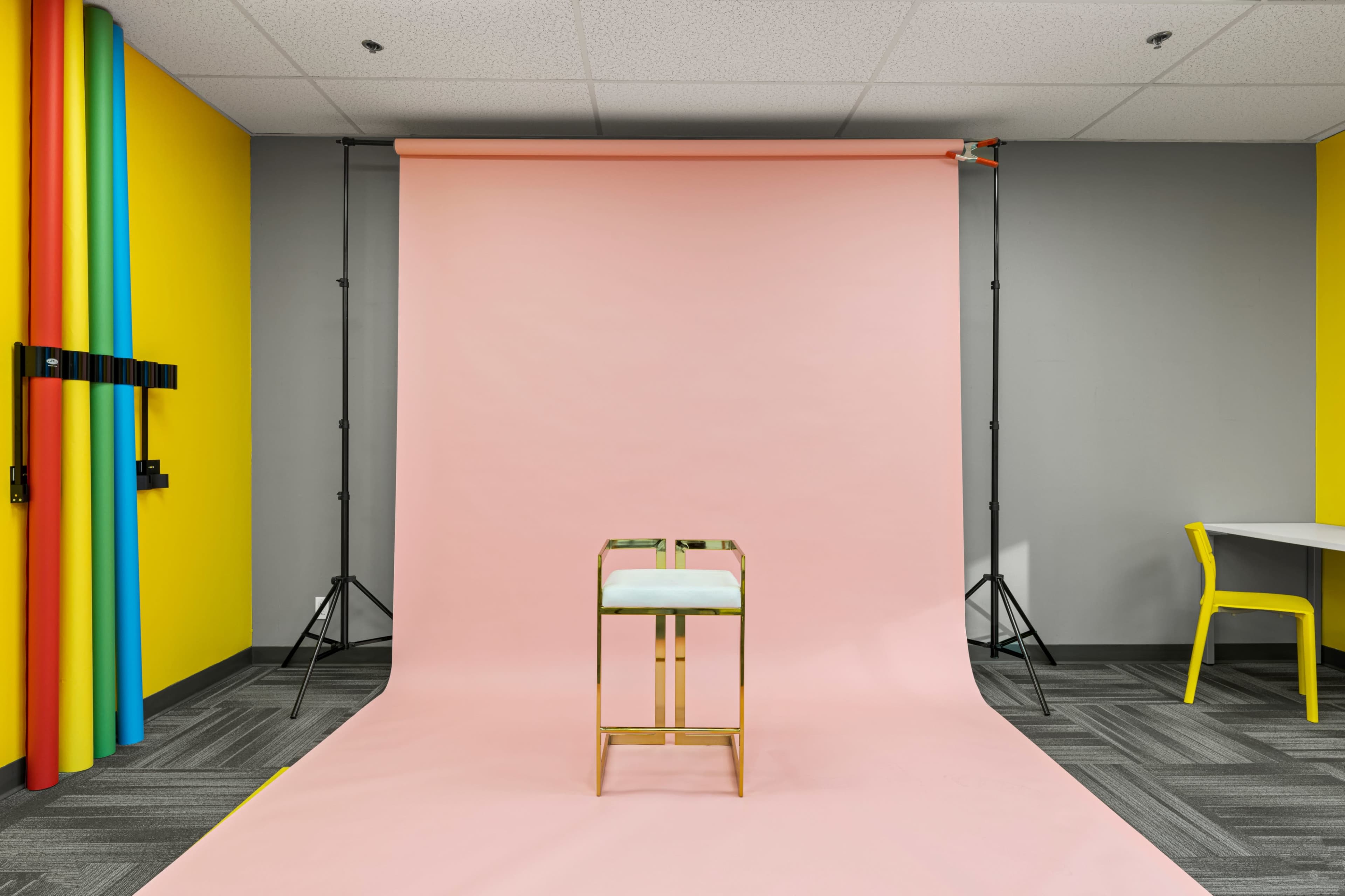 The image shows a photo studio setup with a pink backdrop, a stool at the center, and colorful vertical strips of fabric along one side.