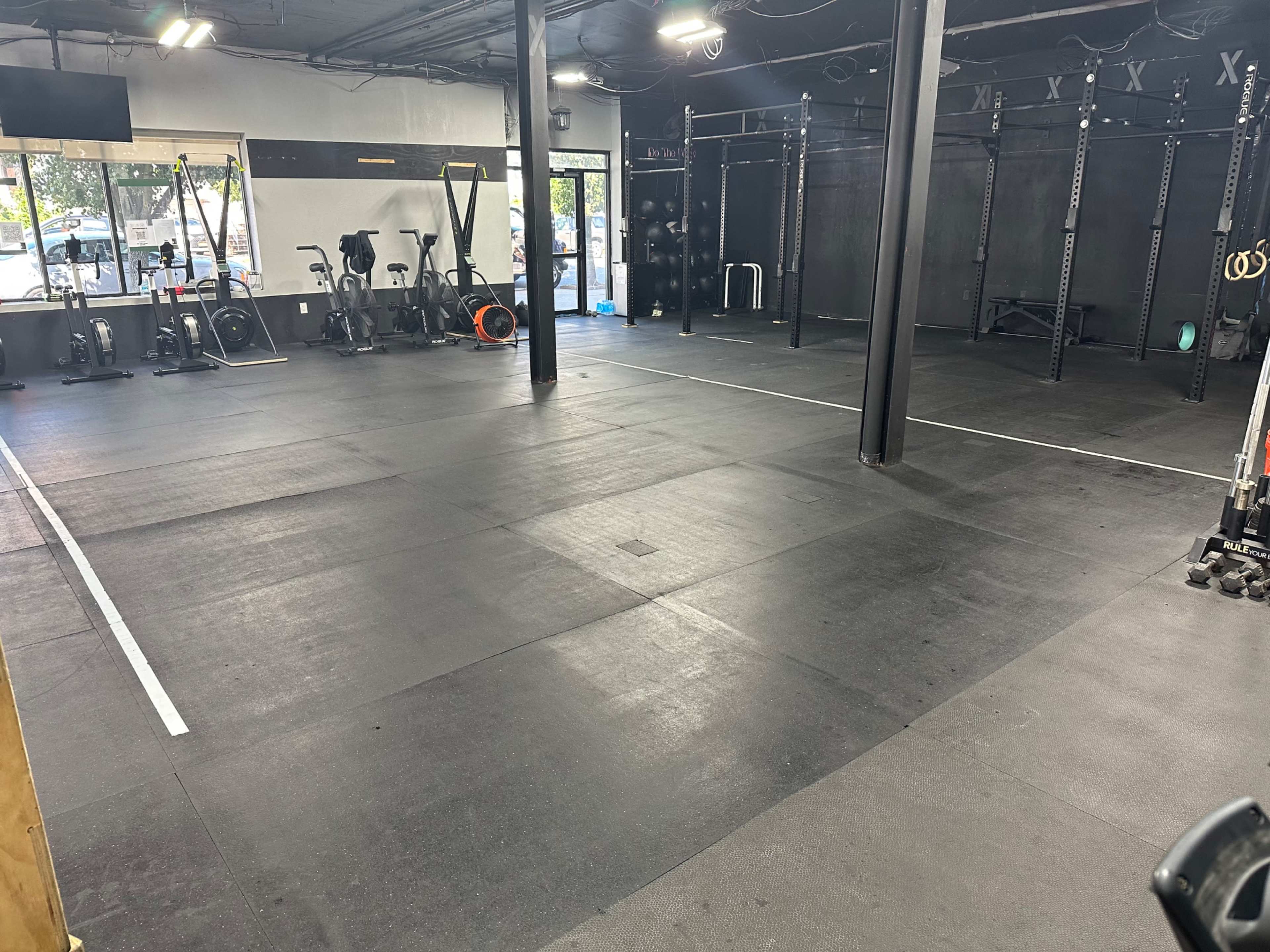 The image shows an empty gym with black rubber flooring, various exercise equipment, and large windows allowing natural light to enter.