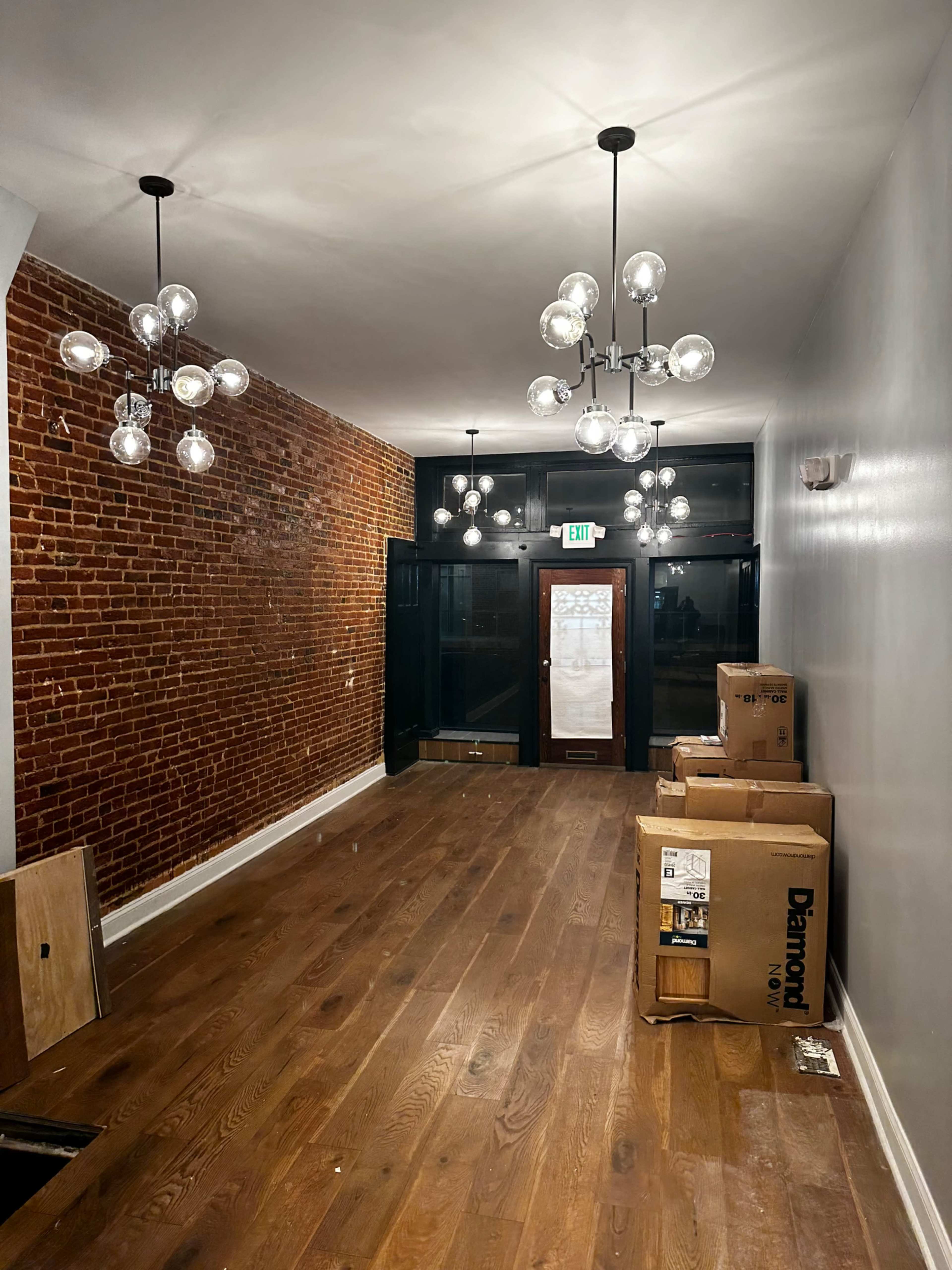 A long, narrow hallway with exposed brick walls, wooden flooring, and multiple decorative light fixtures hanging from the ceiling, leading to a set of glass doors.