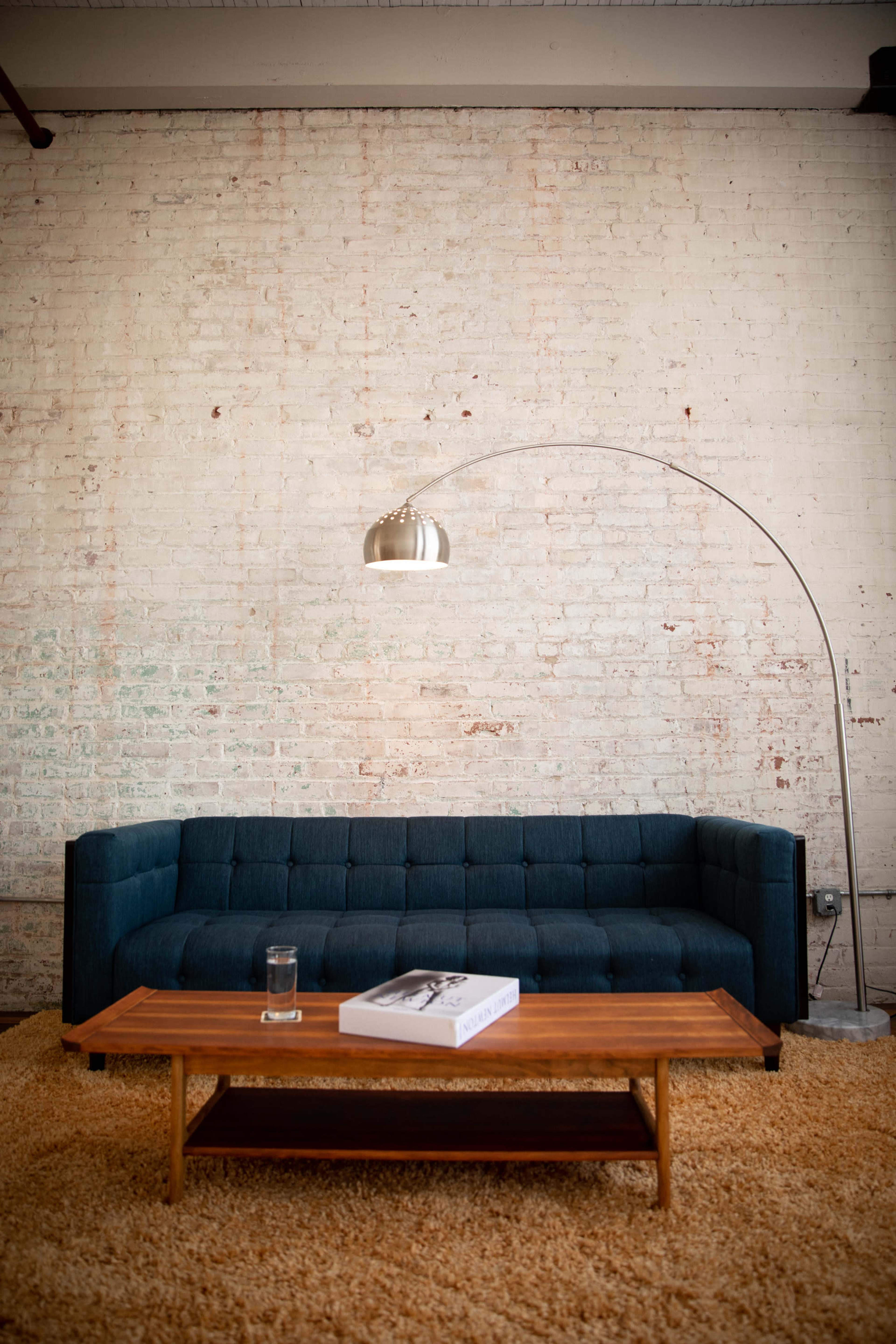 A modern living room featuring a dark blue sofa, a wooden coffee table, and a curved floor lamp, all set against a textured white brick wall.