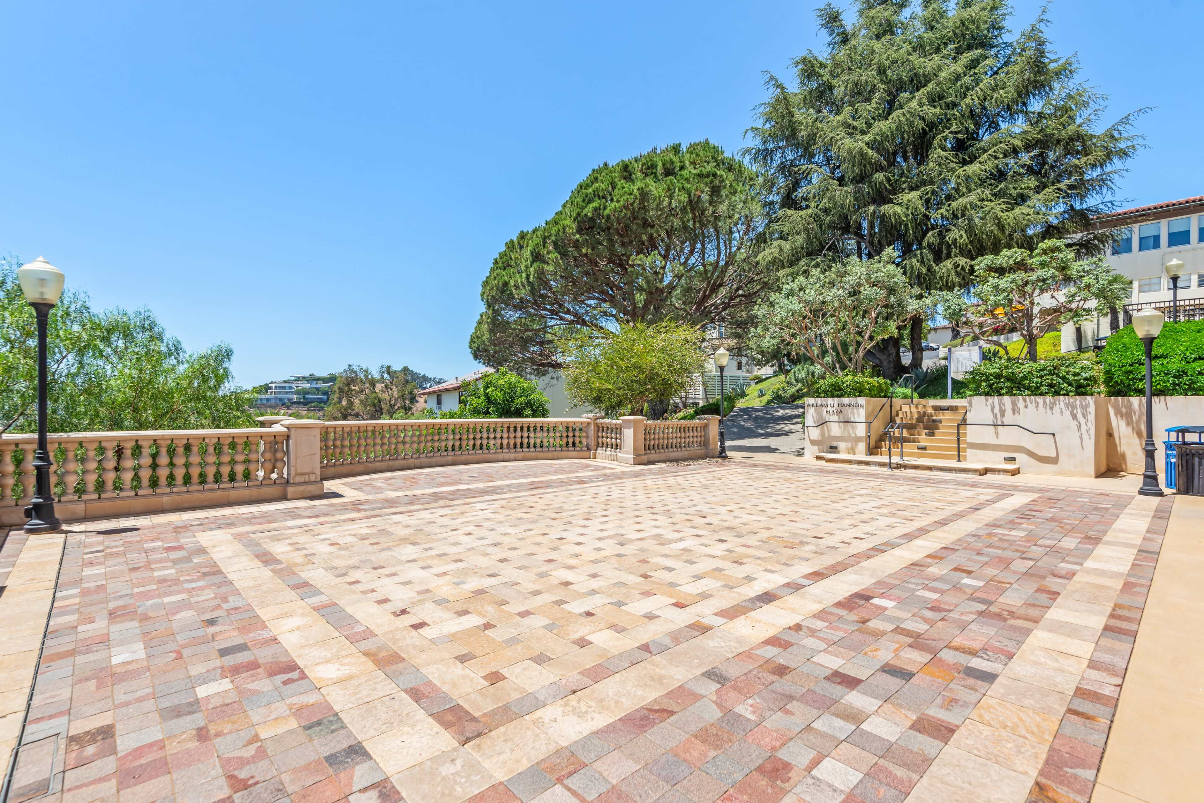 The image shows a spacious terrace with a patterned stone surface, surrounded by trees and buildings in a clear blue sky.