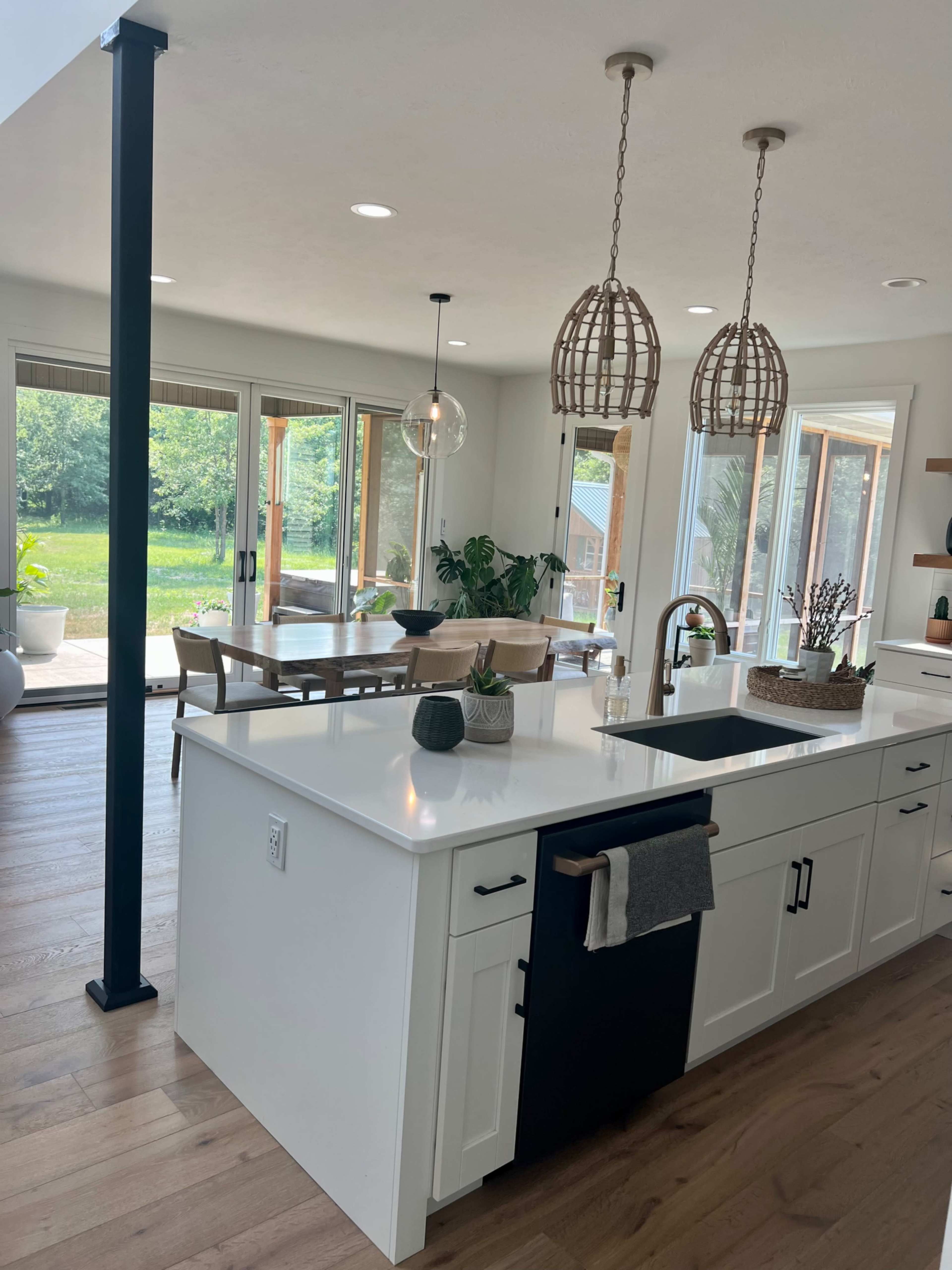 A modern kitchen features a central island with a sink, black cabinetry, and two decorative pendant lights, overlooking a dining area and large windows that provide views of the backyard.