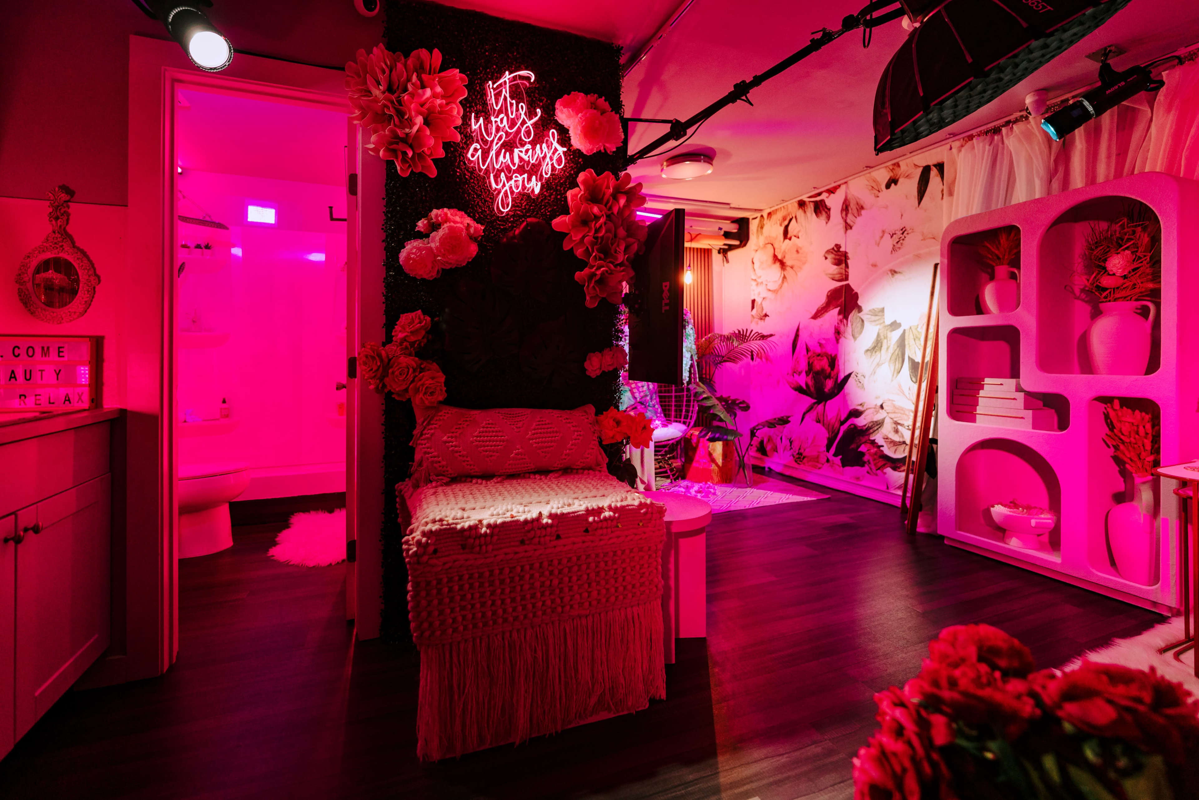 The room features a pink-lit area with floral wall decor, a cozy bed draped in a knitted throw, and a neon sign that reads, "It's you, gorgeous."