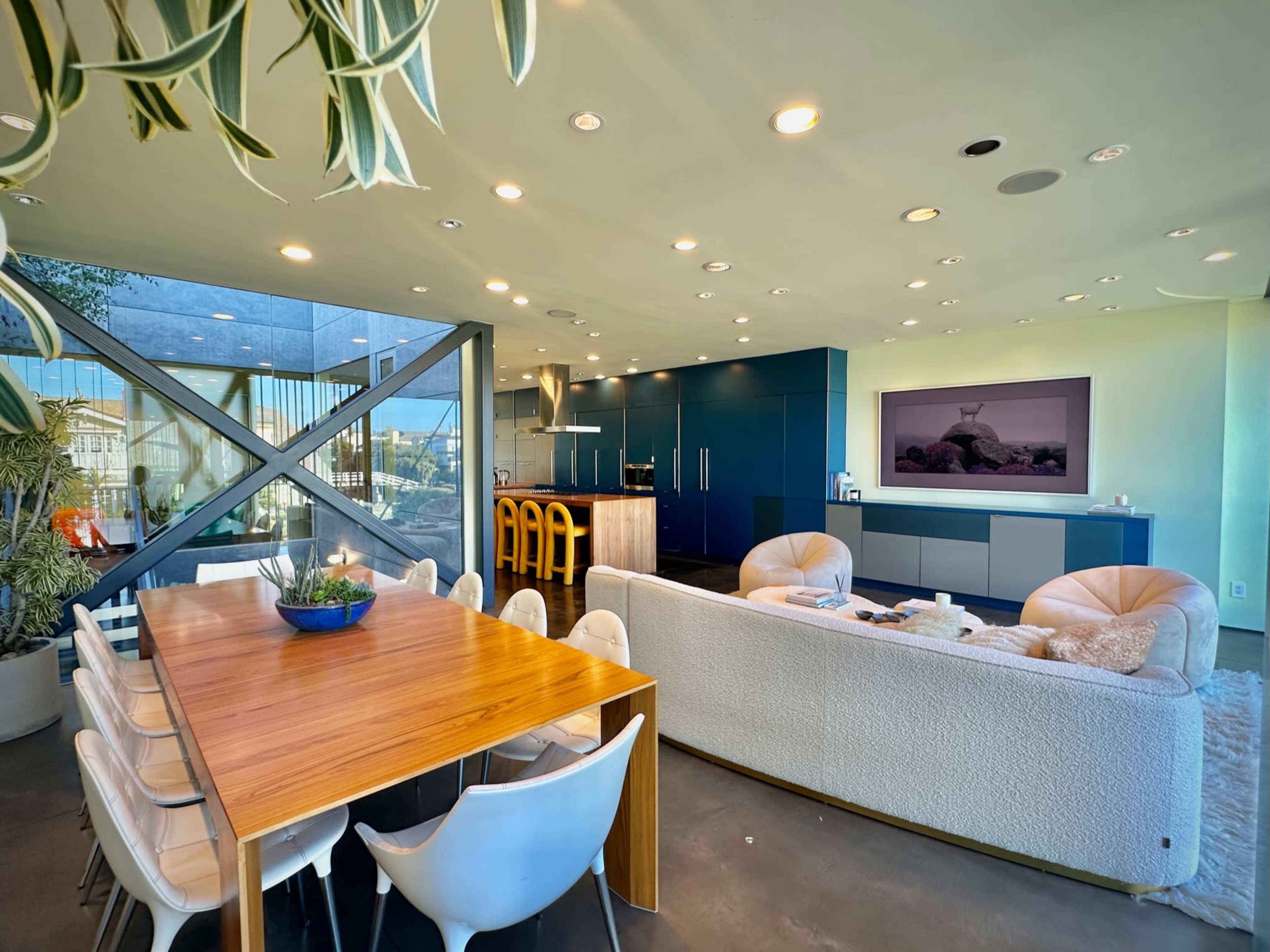 A modern open-plan living space featuring a dining table, a cozy sofa, a kitchen area, and a large television mounted on a wall.