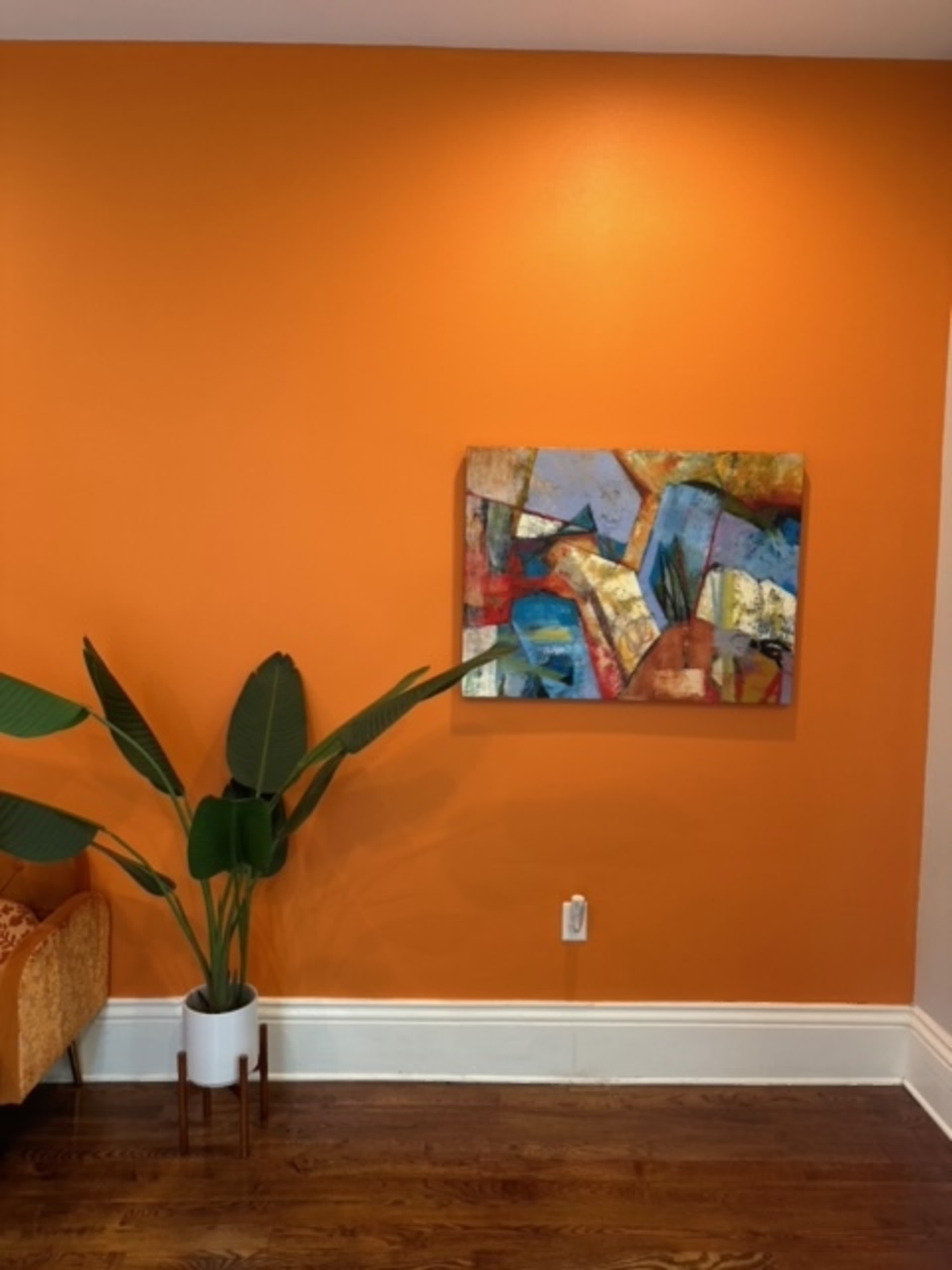The image shows an orange wall with a colorful abstract painting hung on it, accompanied by a green plant in a white pot placed on the floor.