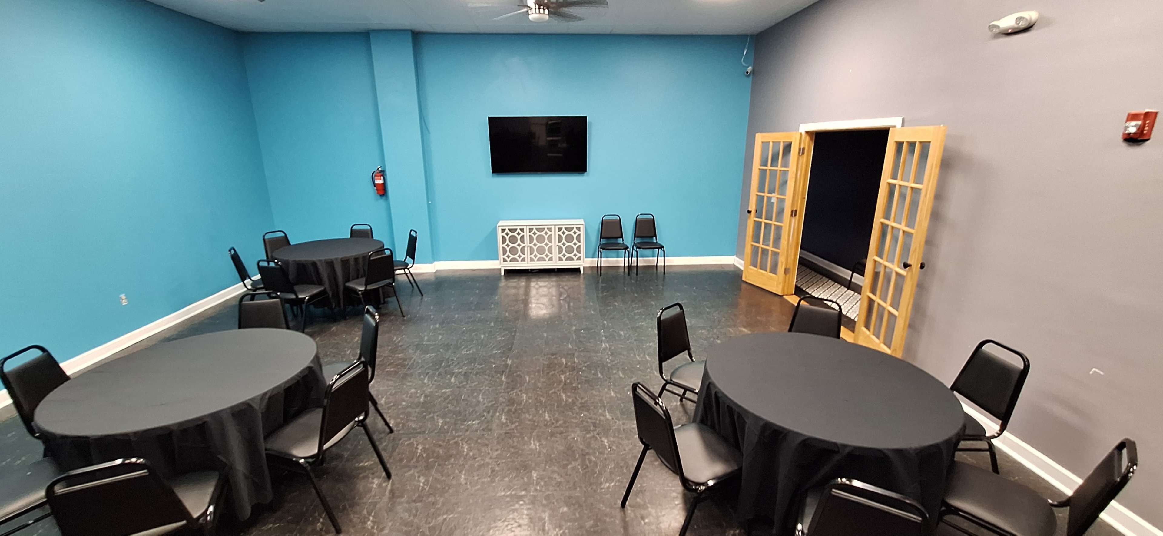 A room with blue walls, black flooring, several round tables covered with black tablecloths, and a television mounted on the wall.