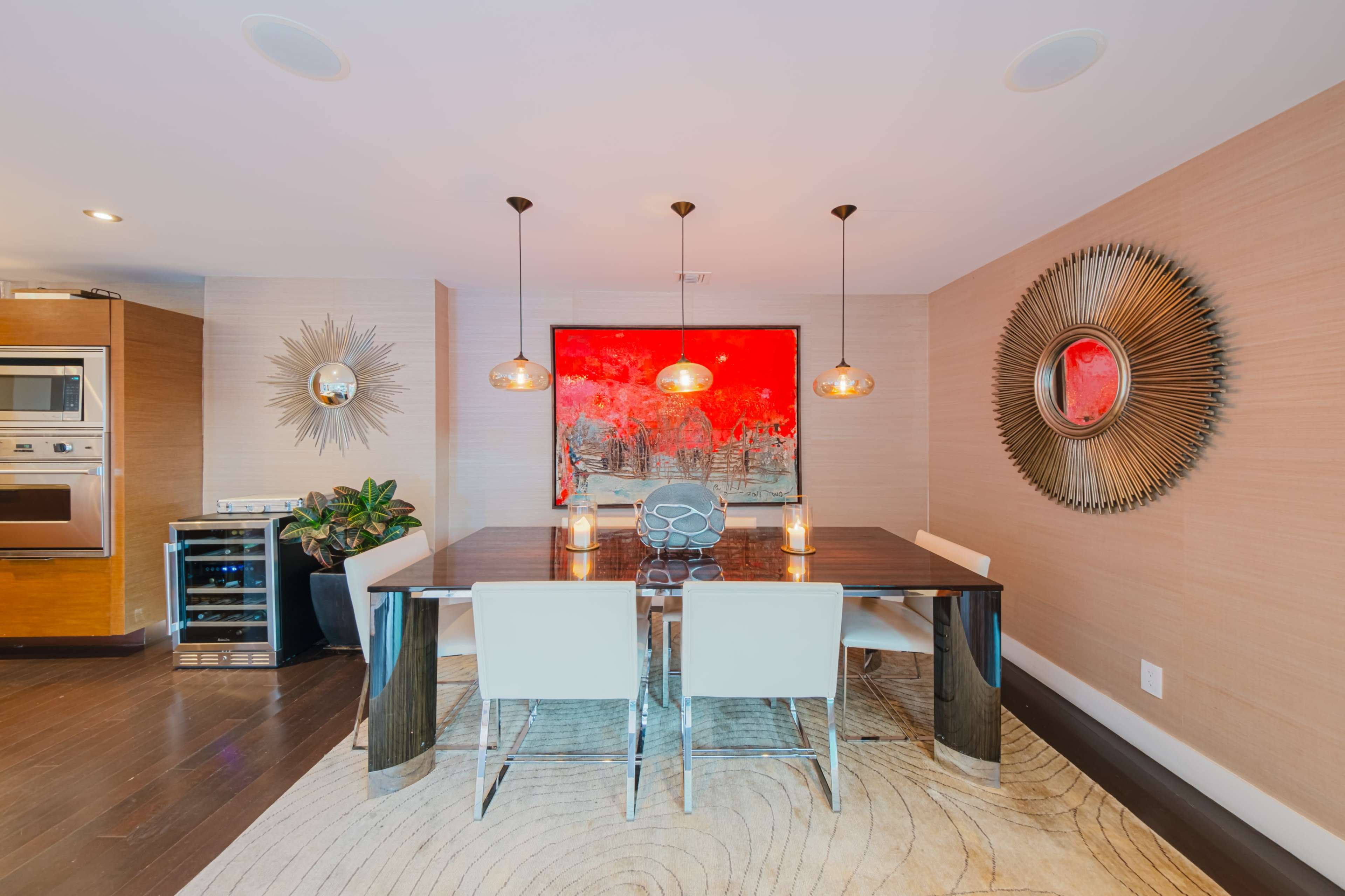 A modern dining area features a sleek table surrounded by white chairs, with a large red artwork on the wall and pendant lights overhead.