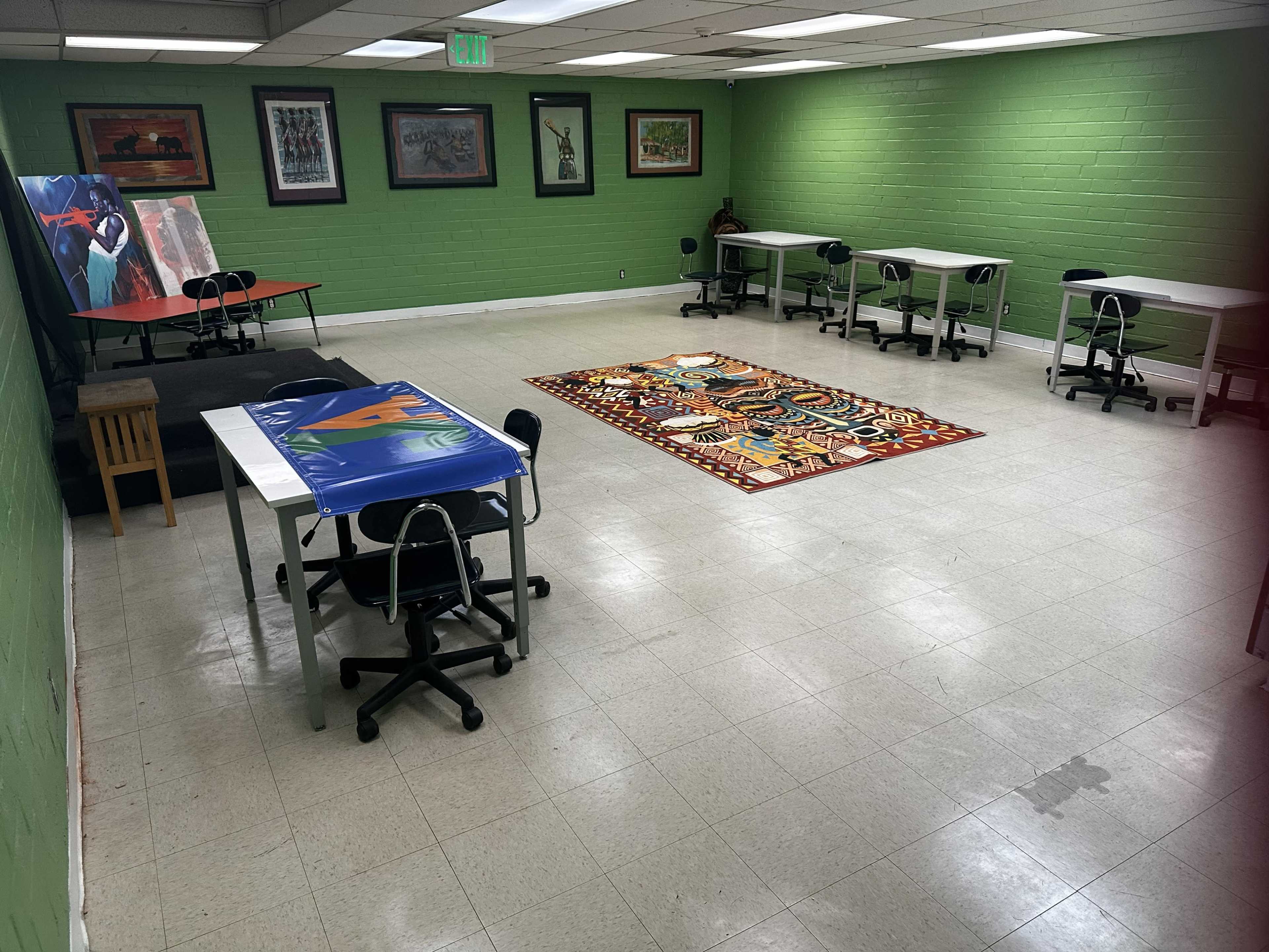 The image shows a room with green walls, multiple tables and chairs arranged in a spacious layout, and a patterned rug in the center.