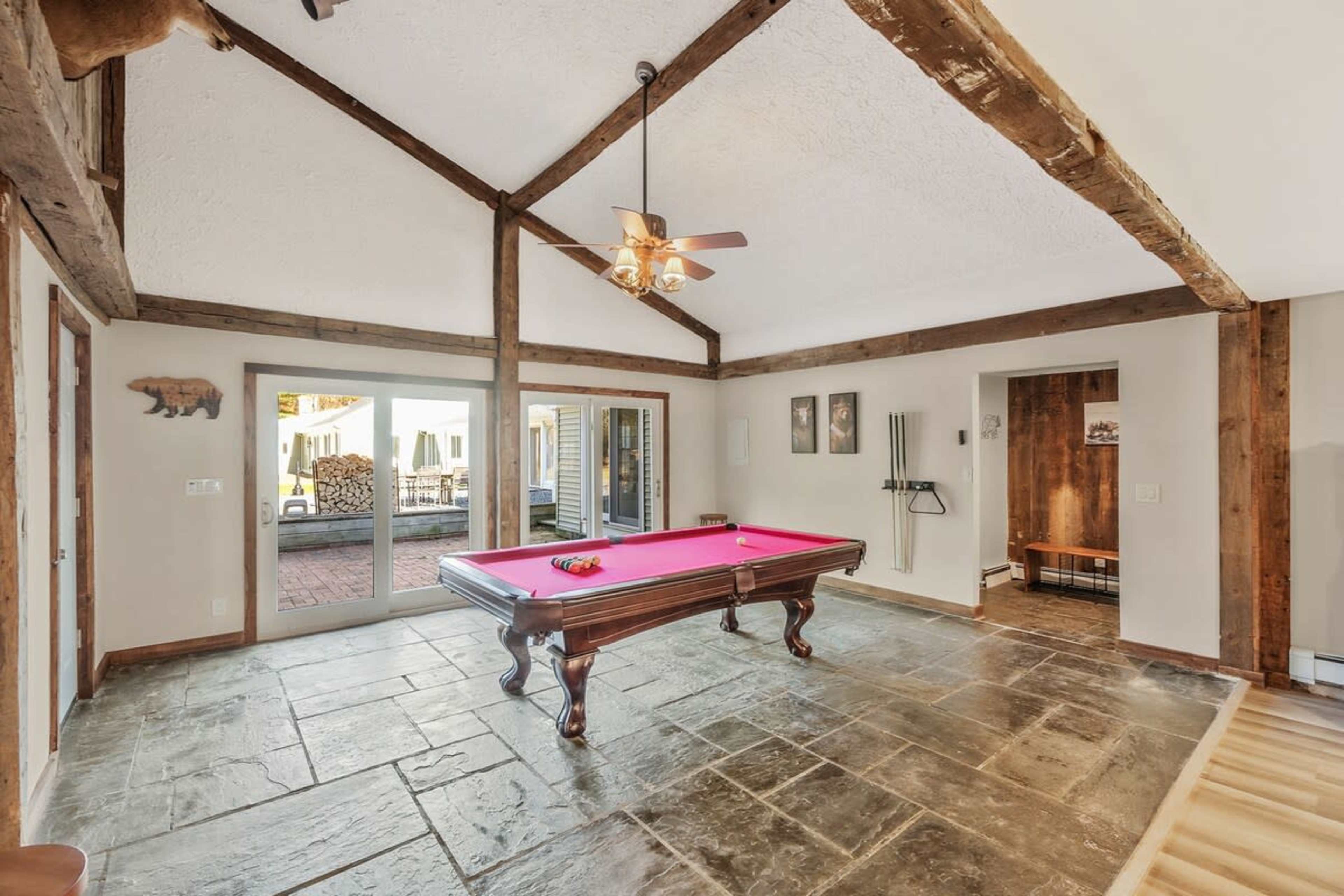 The image shows a game room featuring a pink-felted pool table centered on a slate floor, with exposed wooden beams and large glass doors leading outside.