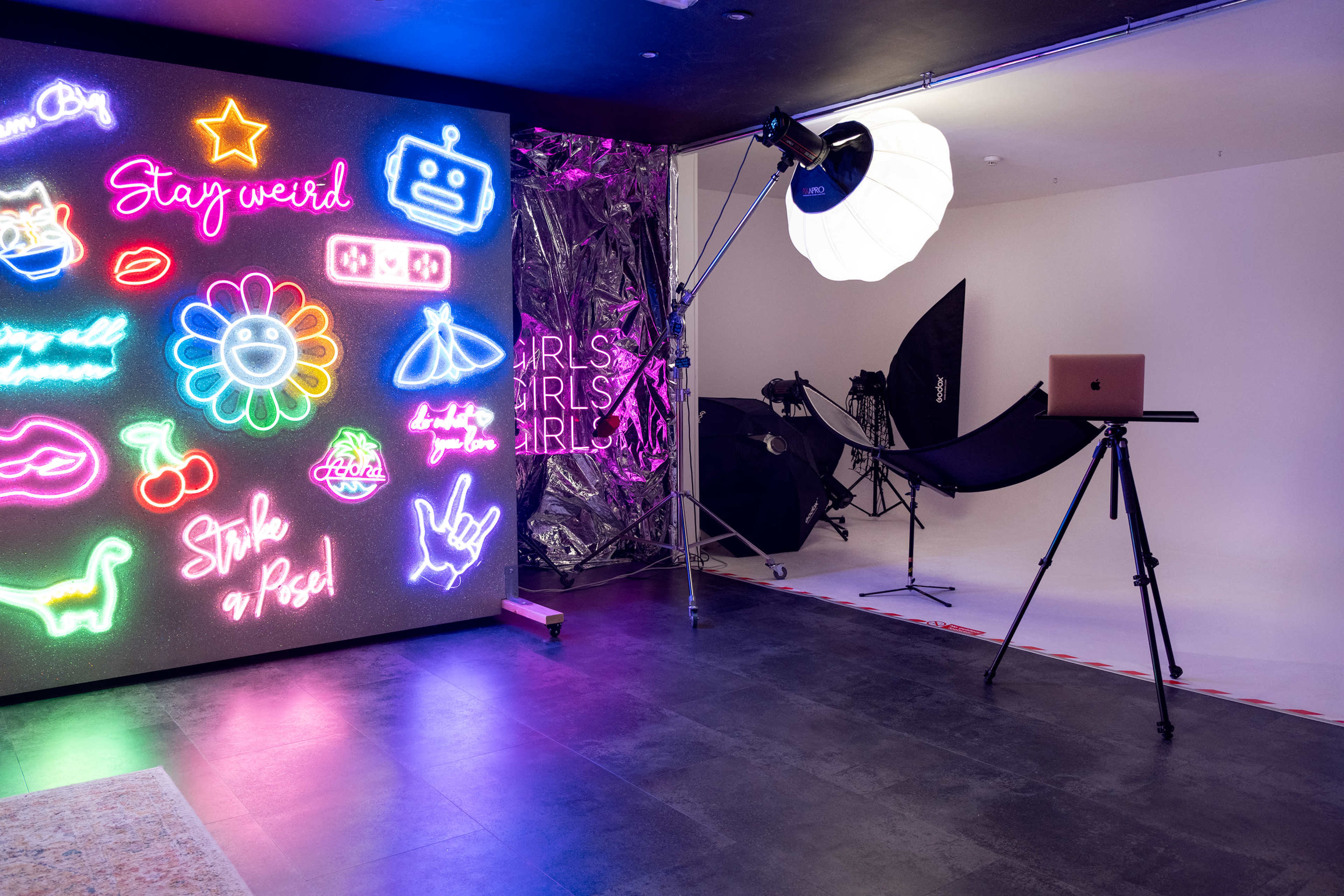 The image shows a photography studio featuring a colorful neon wall with various designs, a lighting setup, and a laptop on a tripod.