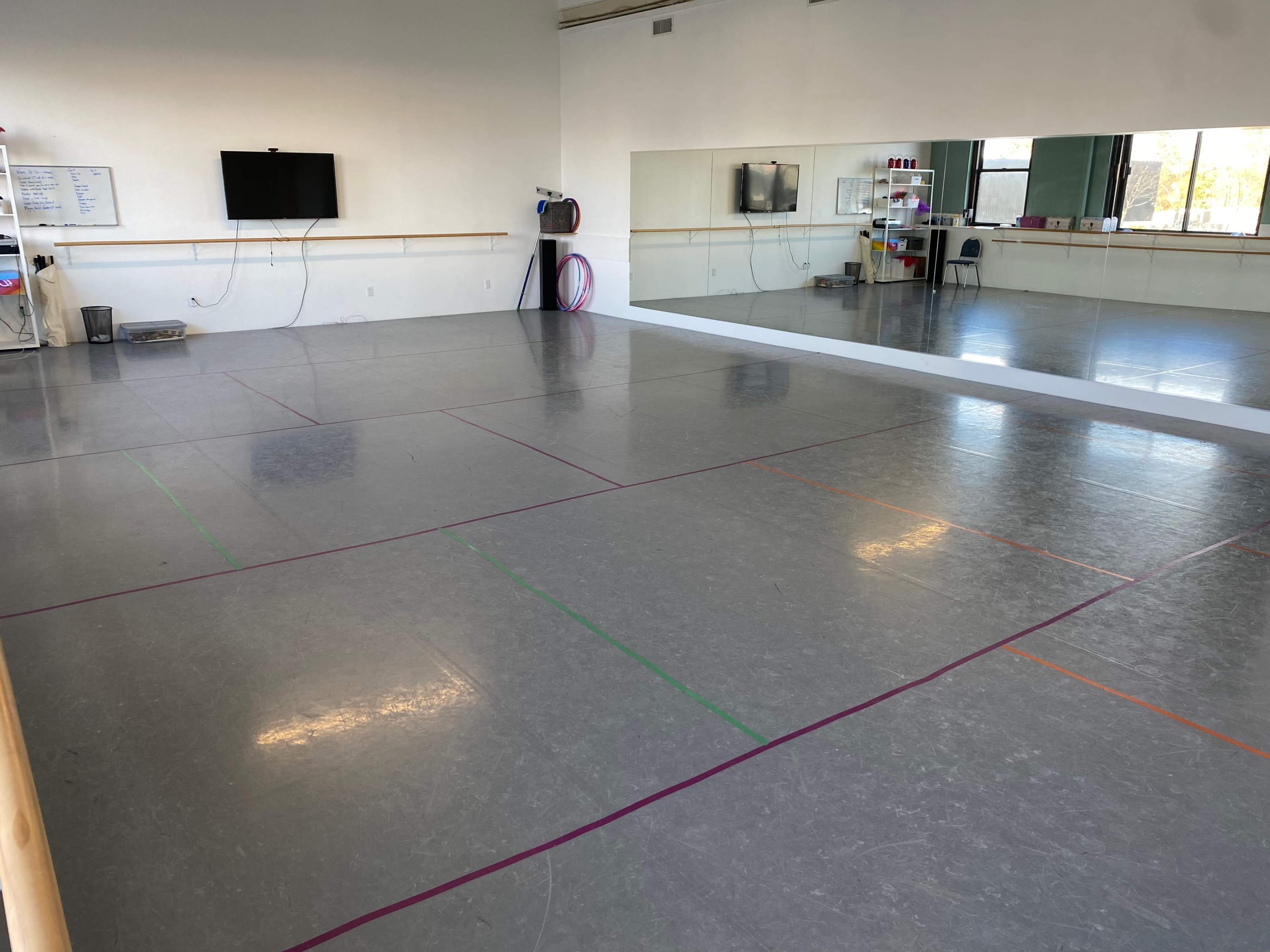 A spacious dance studio with a gray floor, wall mirrors, and minimal decor, featuring marked lines in various colors on the floor.
