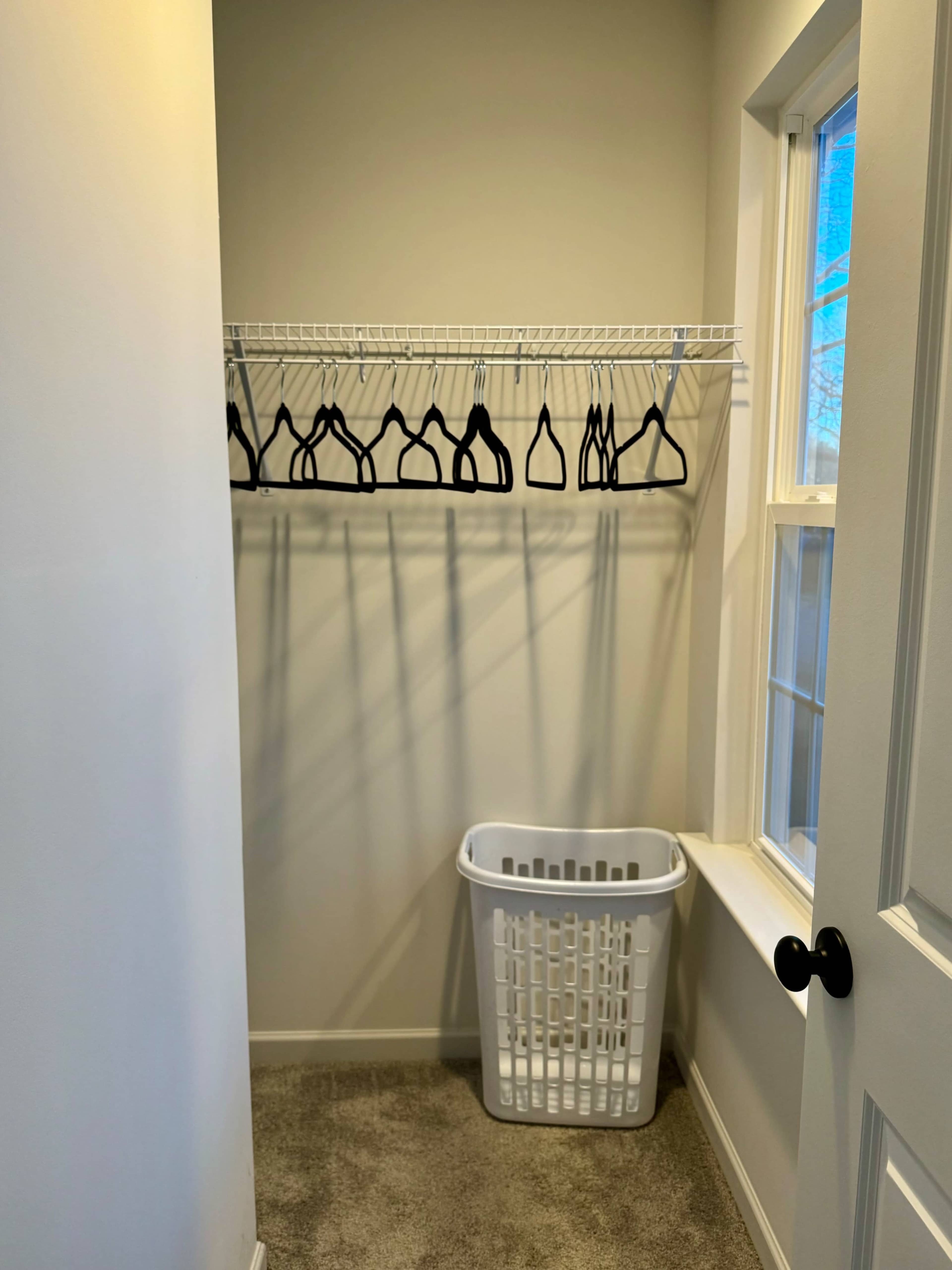 The image shows a small closet with a white wire shelf holding several black hangers and a white laundry basket on the beige carpeted floor next to a window.
